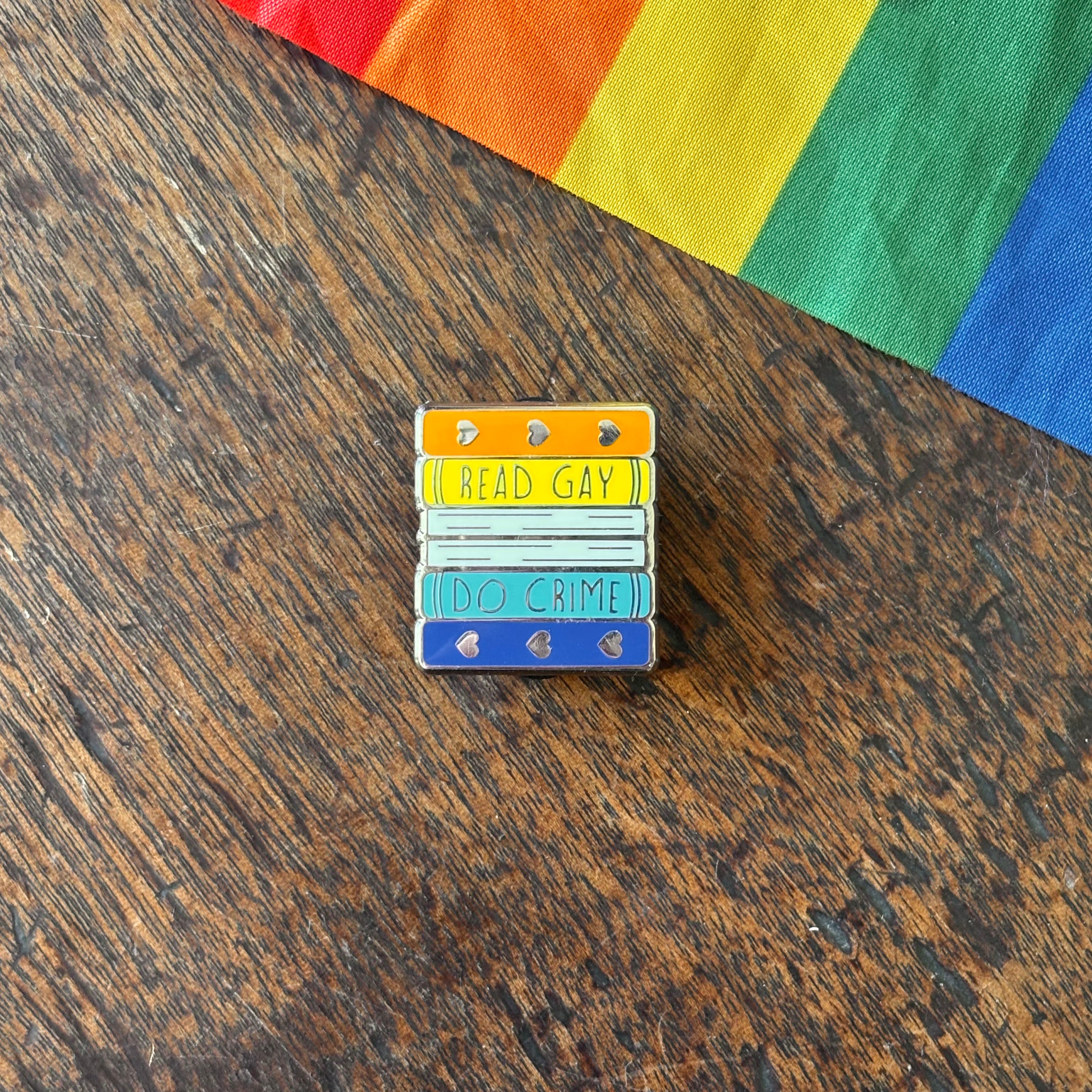 Pin with 'Read Gay, Do Crime' text on a wooden surface with a rainbow fabric background