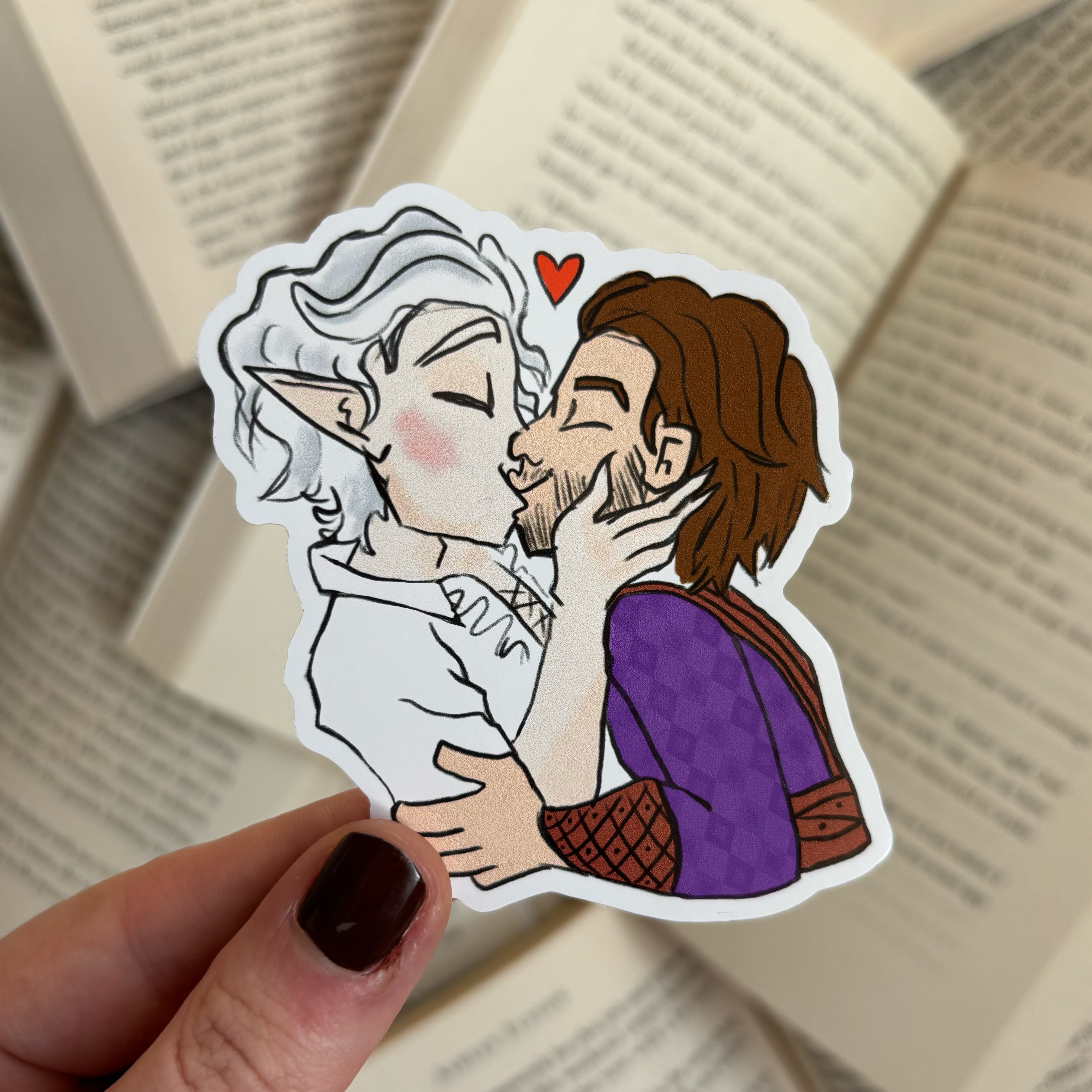 Photo of the Bloodweave inspired sticker featuring Gale and Astarion from Baldur's Gate 3 in an embrace and kissing, held against a background of open books.