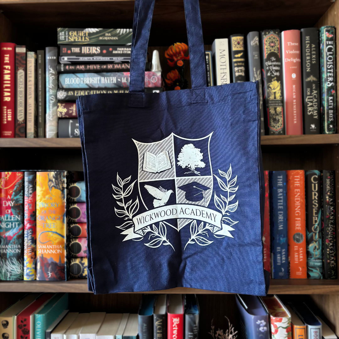 A navy blue canvas tote bag featuring a white printed crest and the words 'WICKWOOD ACADEMY' hanging in front of a wooden bookshelf filled with books.