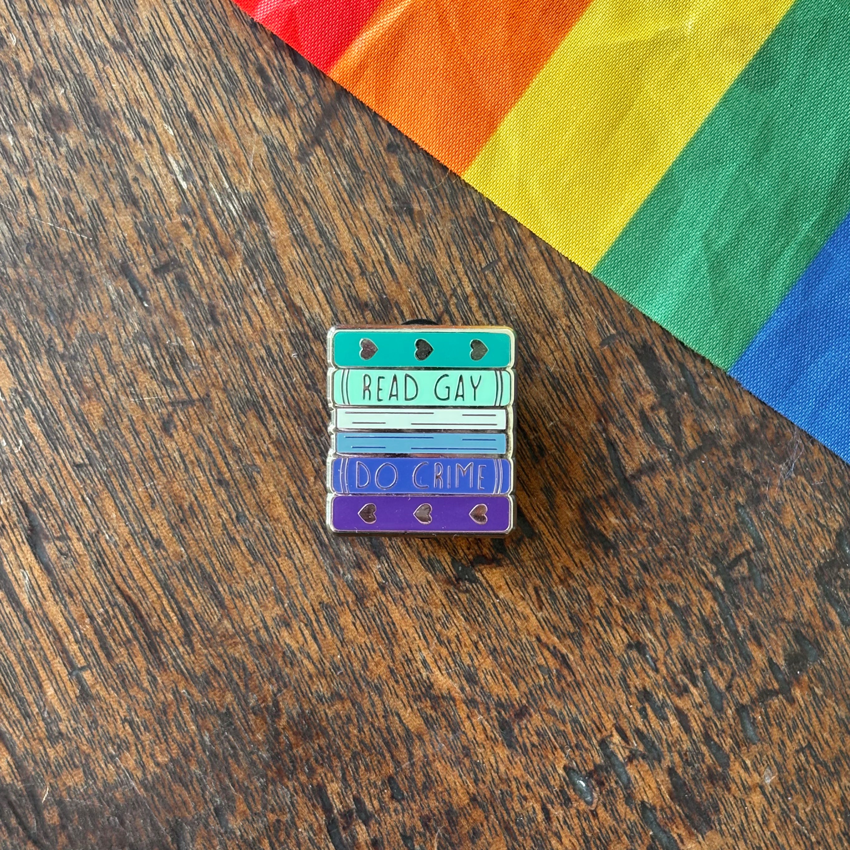 Pin with 'Read Gay, Do Crime' text on a wooden surface with a rainbow fabric background