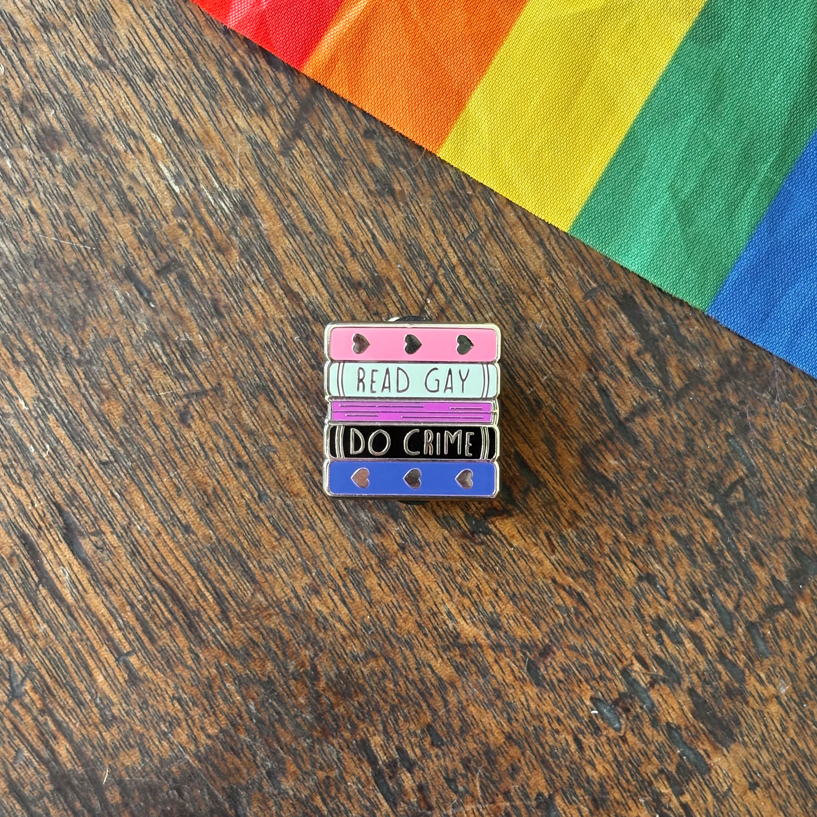 Pin with 'Read Gay, Do Crime' text on a wooden surface with a rainbow fabric background