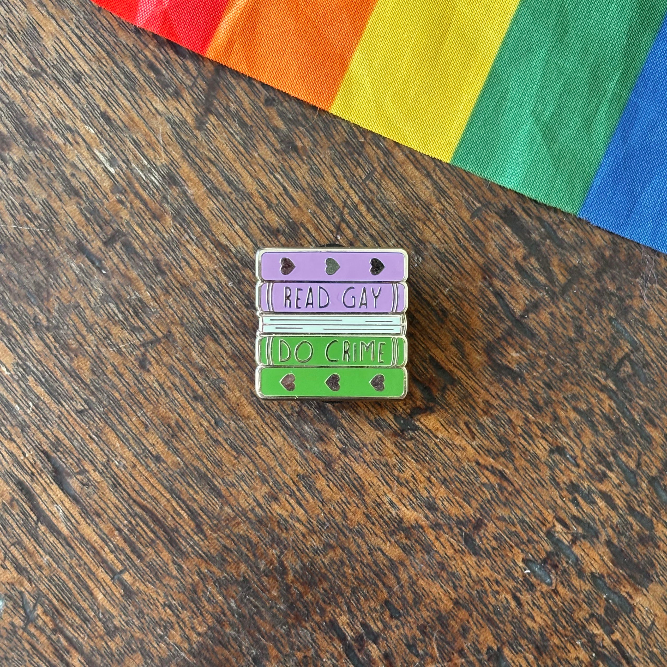 Pin with 'Read Gay, Do Crime' text on a wooden surface with a rainbow fabric background