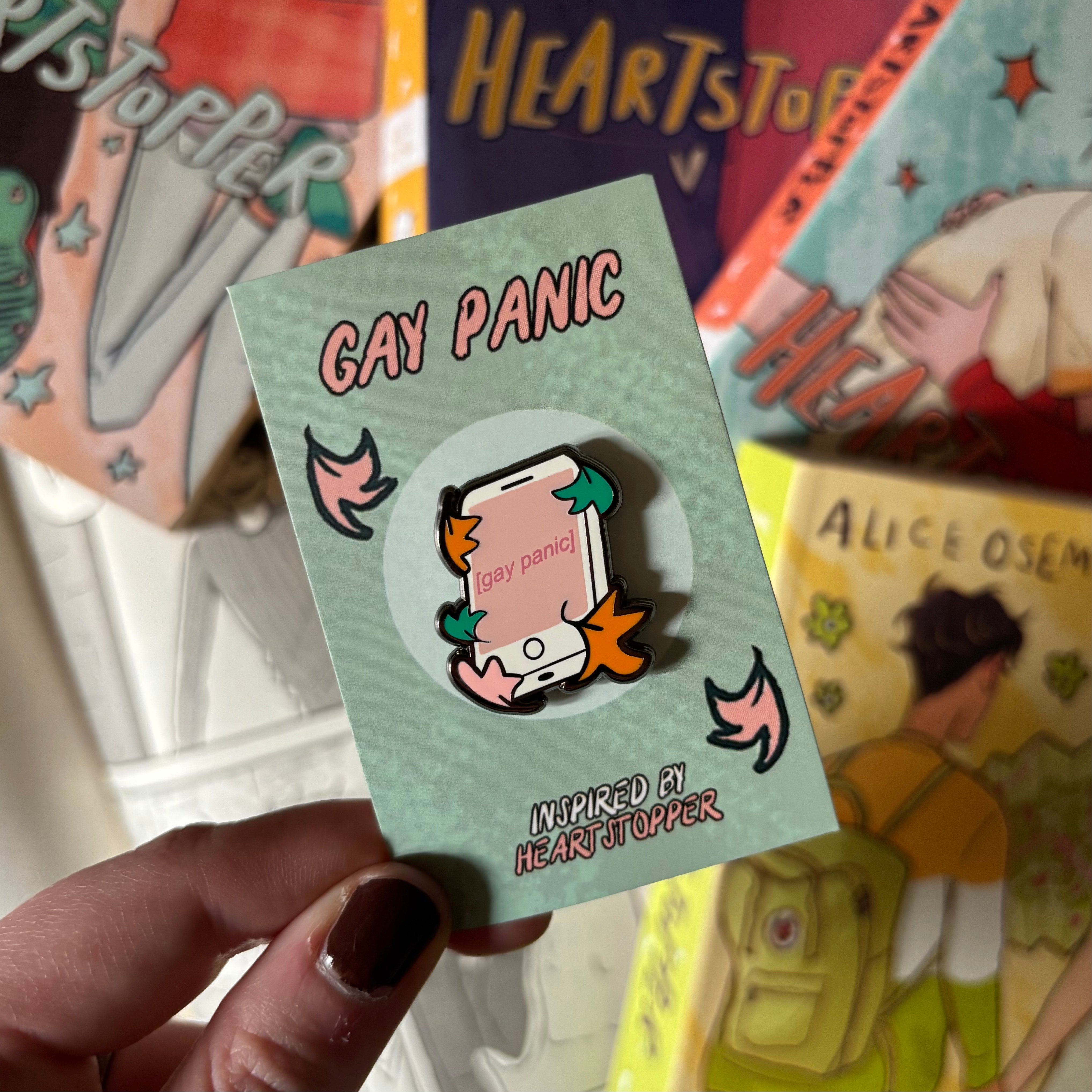 Heartstopper inspired enamel pin with 'Gay Panic' design, held in front of the Heartstopper graphic novels by Alice Oseman.