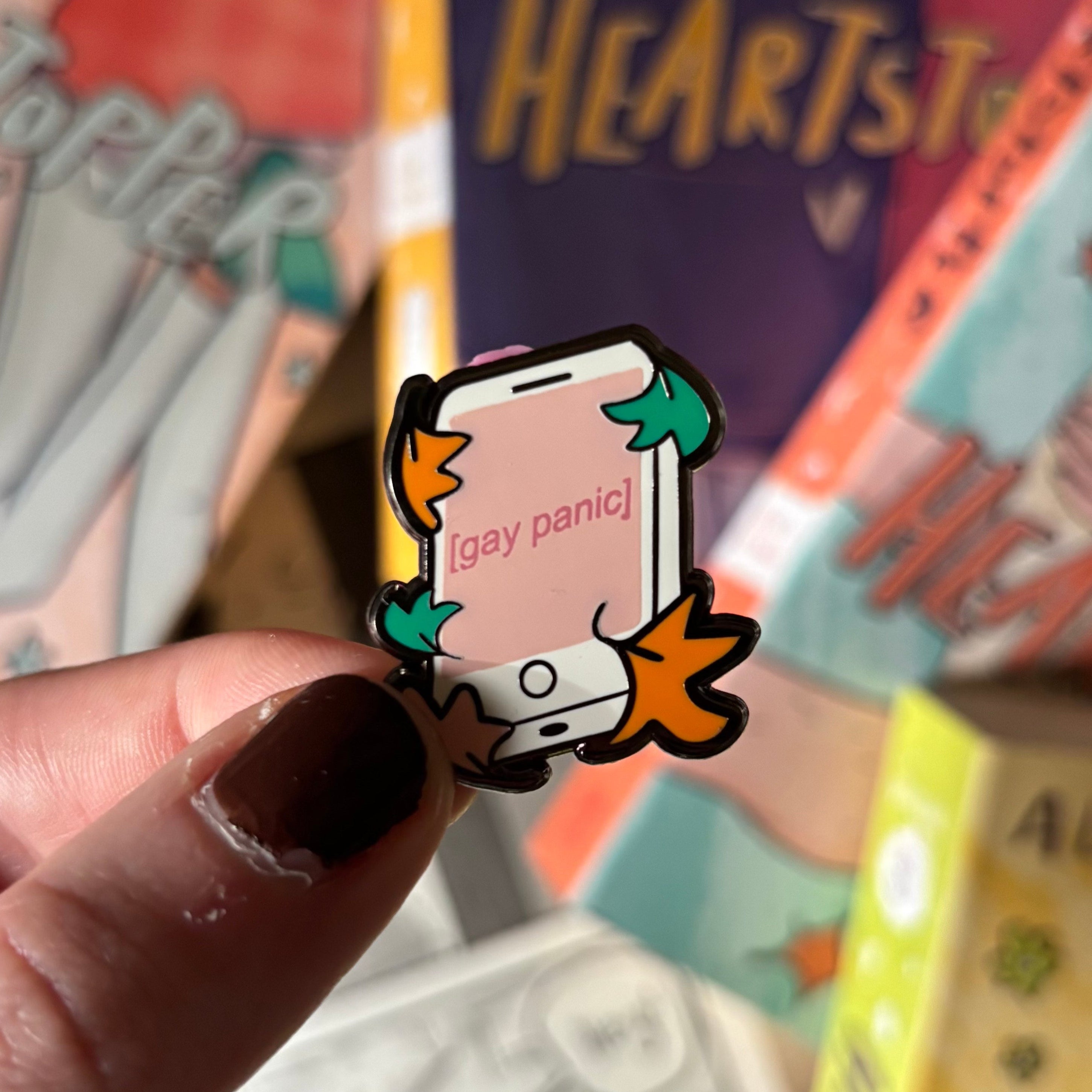 Heartstopper inspired enamel pin with 'Gay Panic' design, held in front of the Heartstopper graphic novels by Alice Oseman.