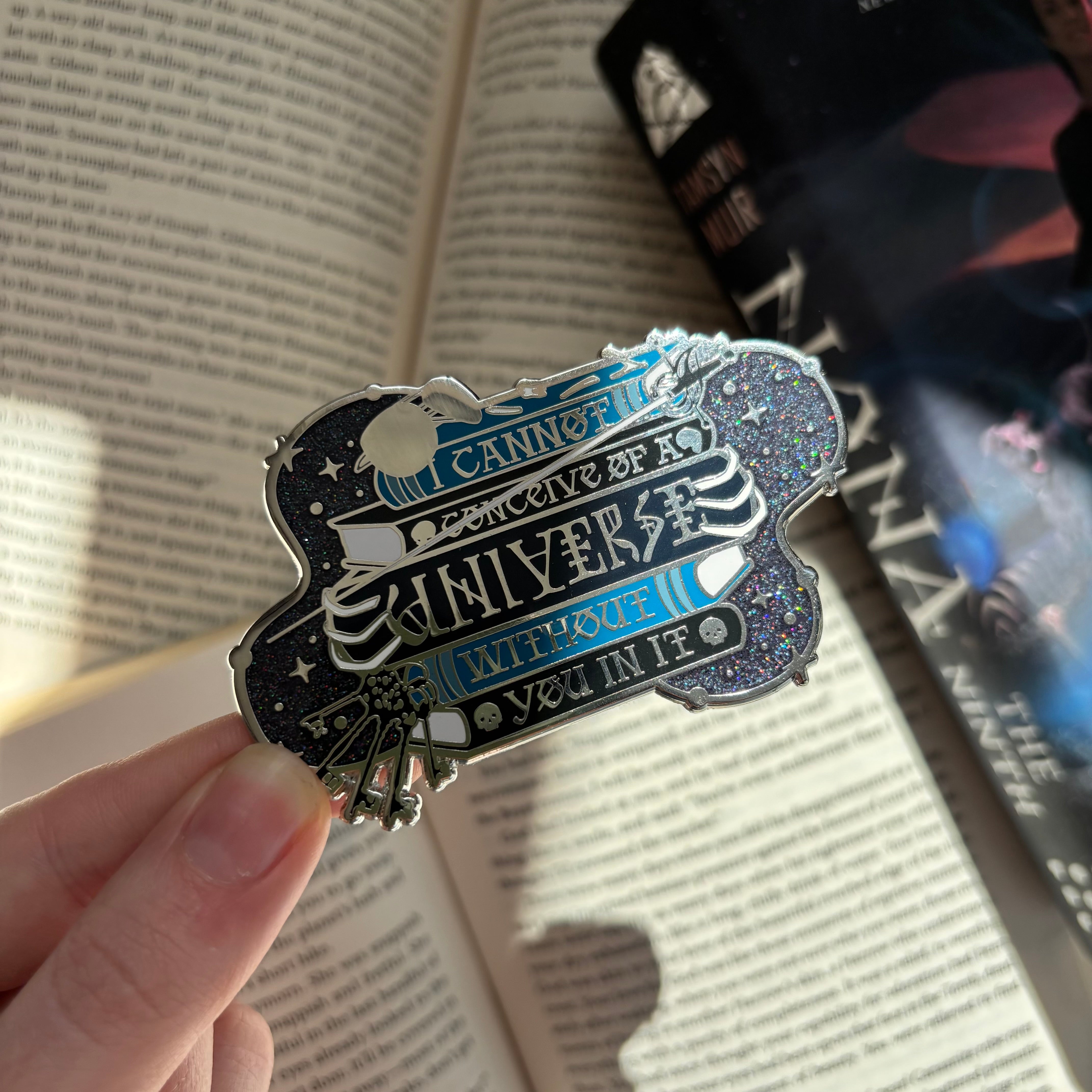 A metallic enamel pin inspired by Gideon the Ninth by Tamsyn Muir, being held over an open book. text on the enamel pin quote: I cannot conceive of a universe without you in it.