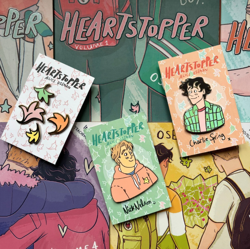 A bundle of our officially licensed Heartstopper pins. Features Nick Nelson, Charlie Sprint, and mini leaf pins, all designed by Alice Oseman. Pictured against a background of Heartstopper graphic novels.