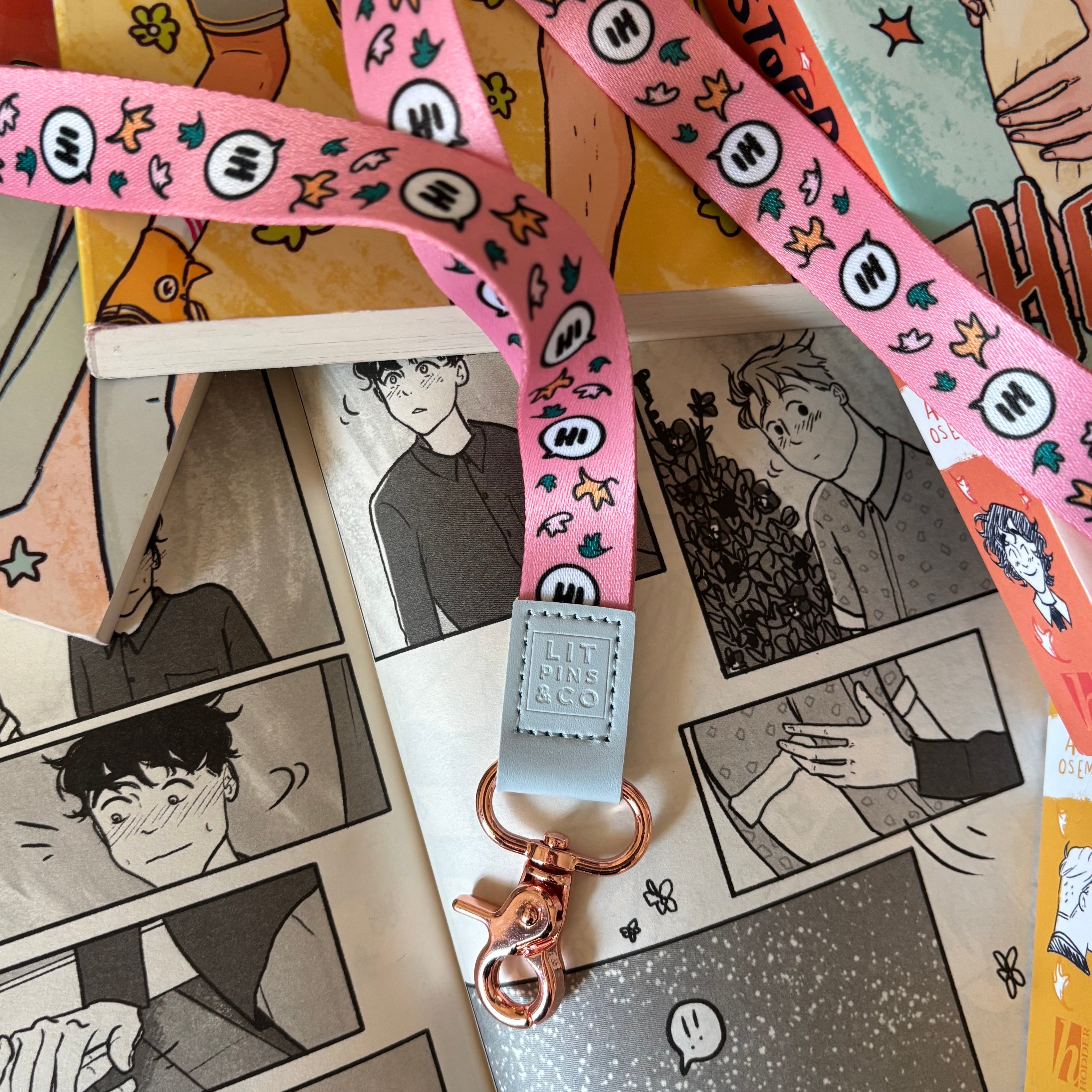 Pink lanyard inspired by Heartstopper by Alice Oseman. Decorated with cartoon speech bubbles reading "Hi" and leaves. Pictured against a background of the Heartstopper graphic novels.
