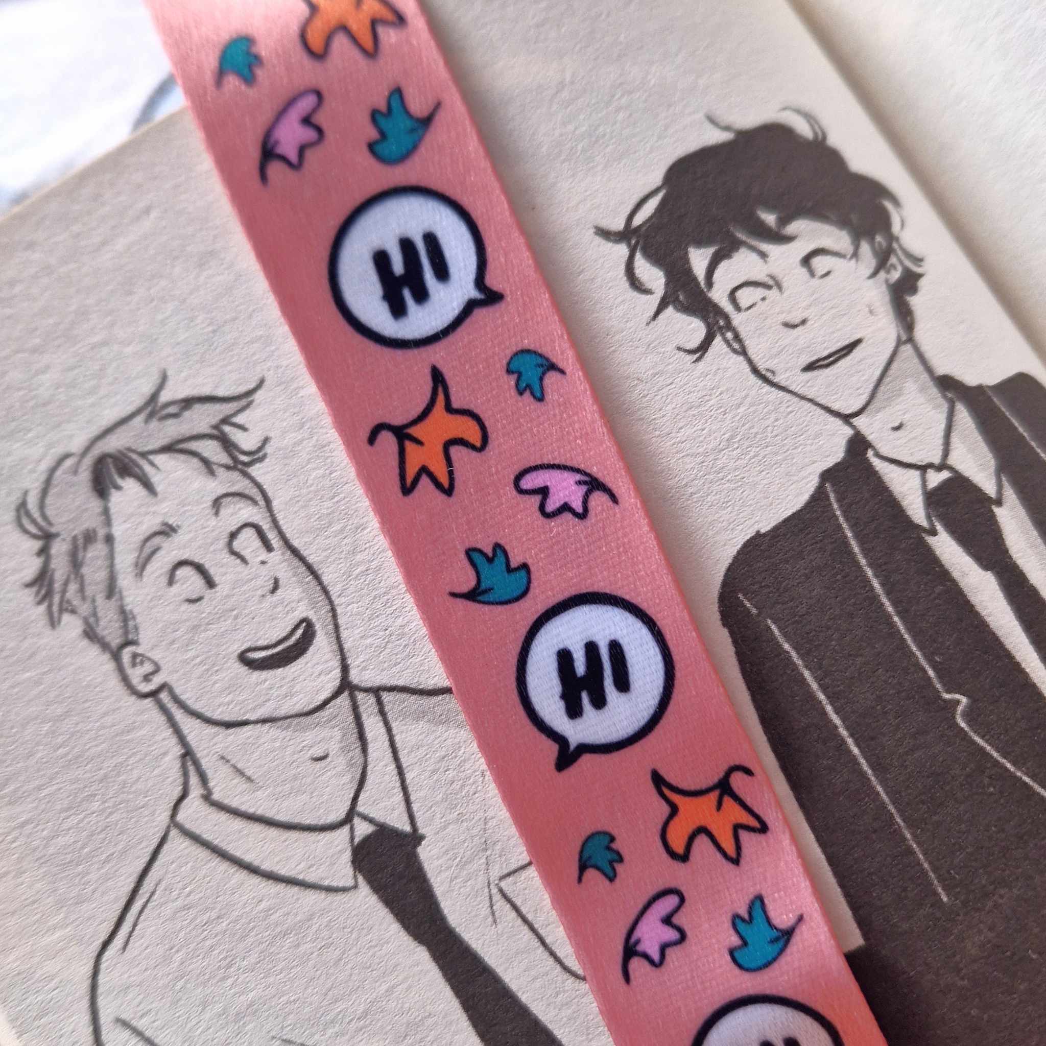 Pink lanyard inspired by Heartstopper by Alice Oseman. Decorated with cartoon speech bubbles reading "Hi" and leaves. Pictured against a background of the Heartstopper graphic novels.