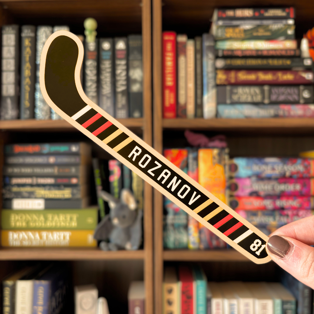A black bookmark shaped like a hockey stick with text reading "Rozanov". The bookmark is held in front of a blurred bookshelf