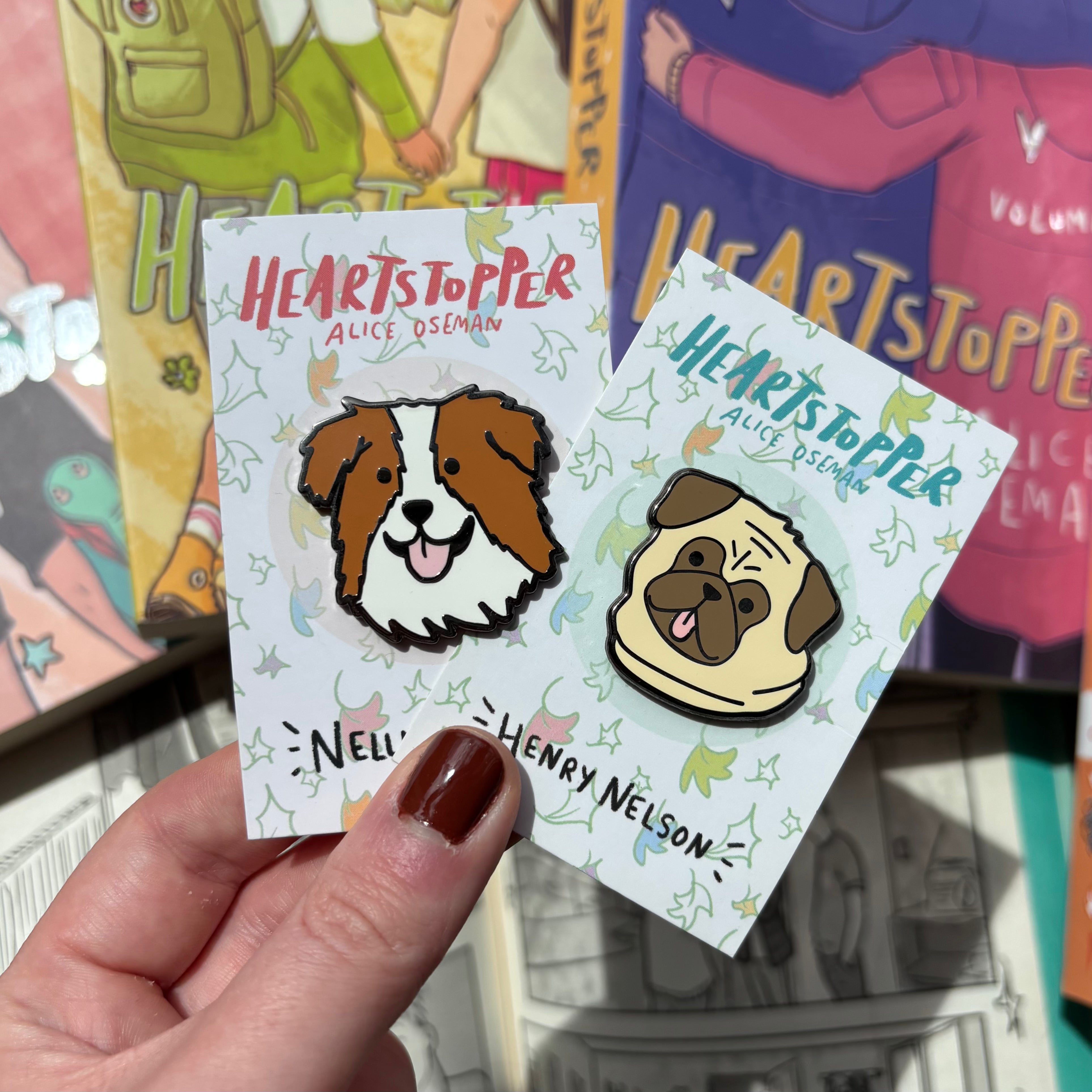 Two officially licensed Heartstopper pins featuring Nellie and Henry the dogs, held against a background of blurred Heartstopper books.