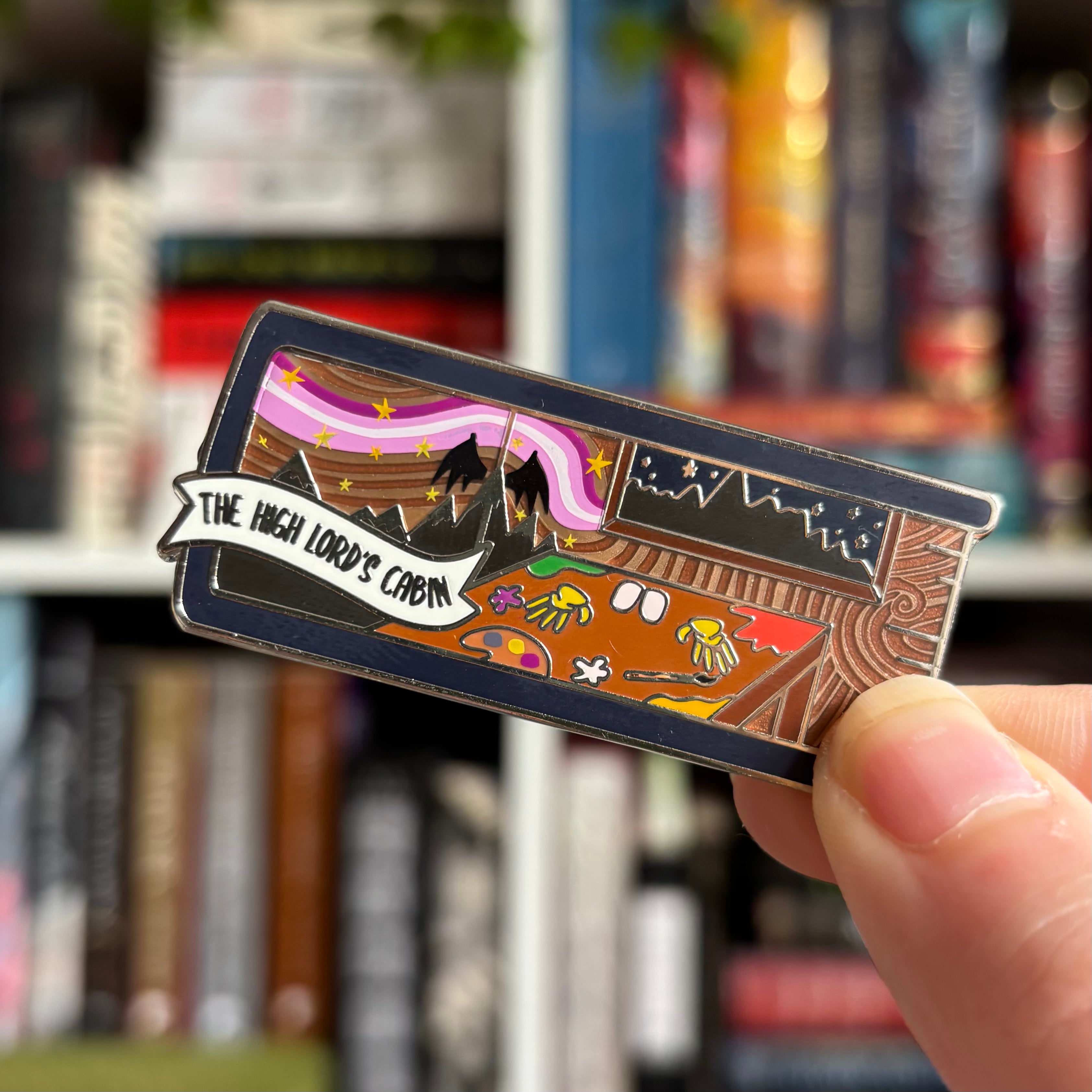 The High Lord's Cabin build your own book stack enamel pin inspired by Sarah J Maas A Court of Throne and Roses series, held against a bookshelf