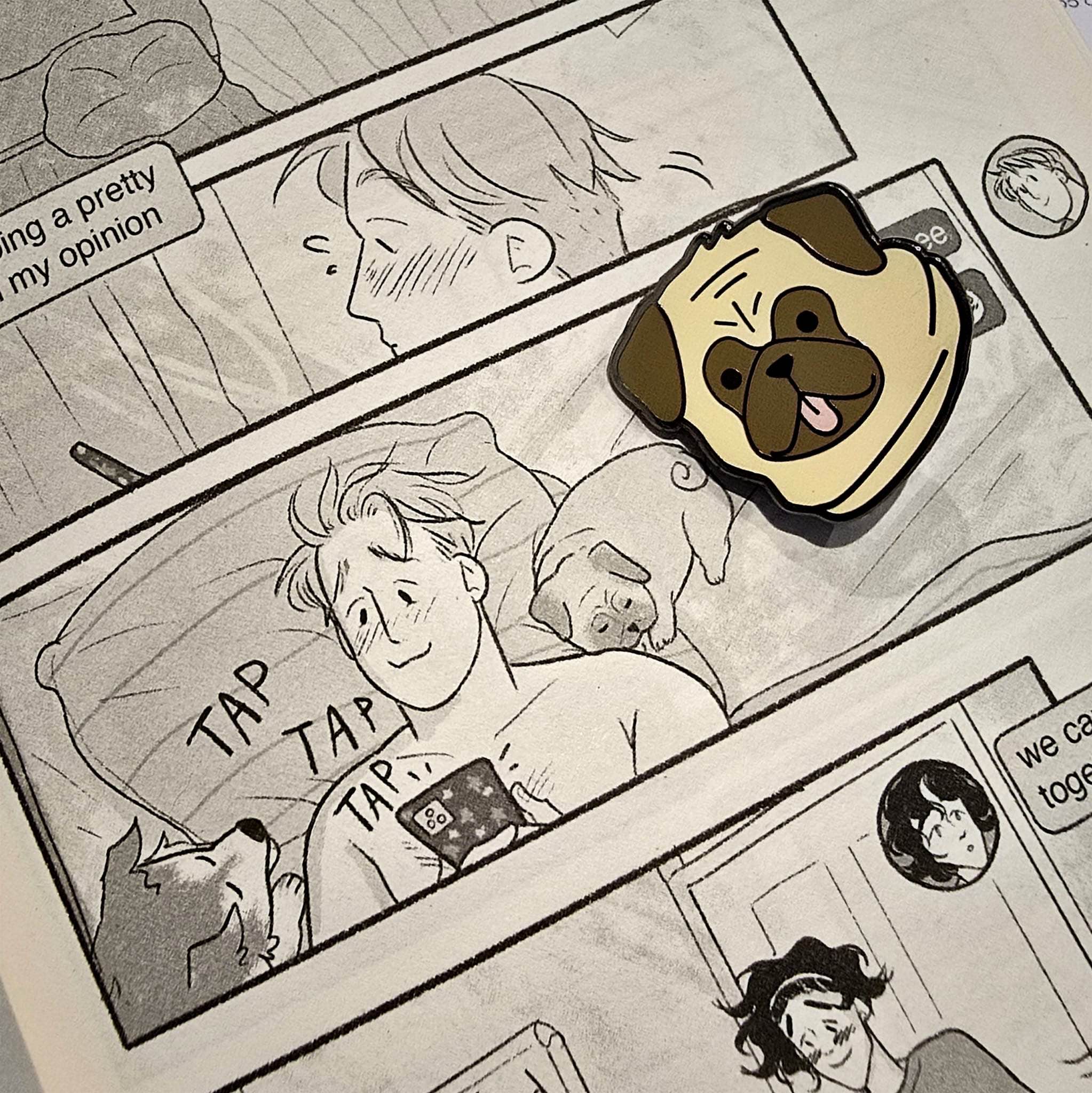 The Henry Nelson enamel pin ressting on the open pages of the Heartstopper graphic novel on the page which Nick is lying in bed with Nellie and Henry on his phone.