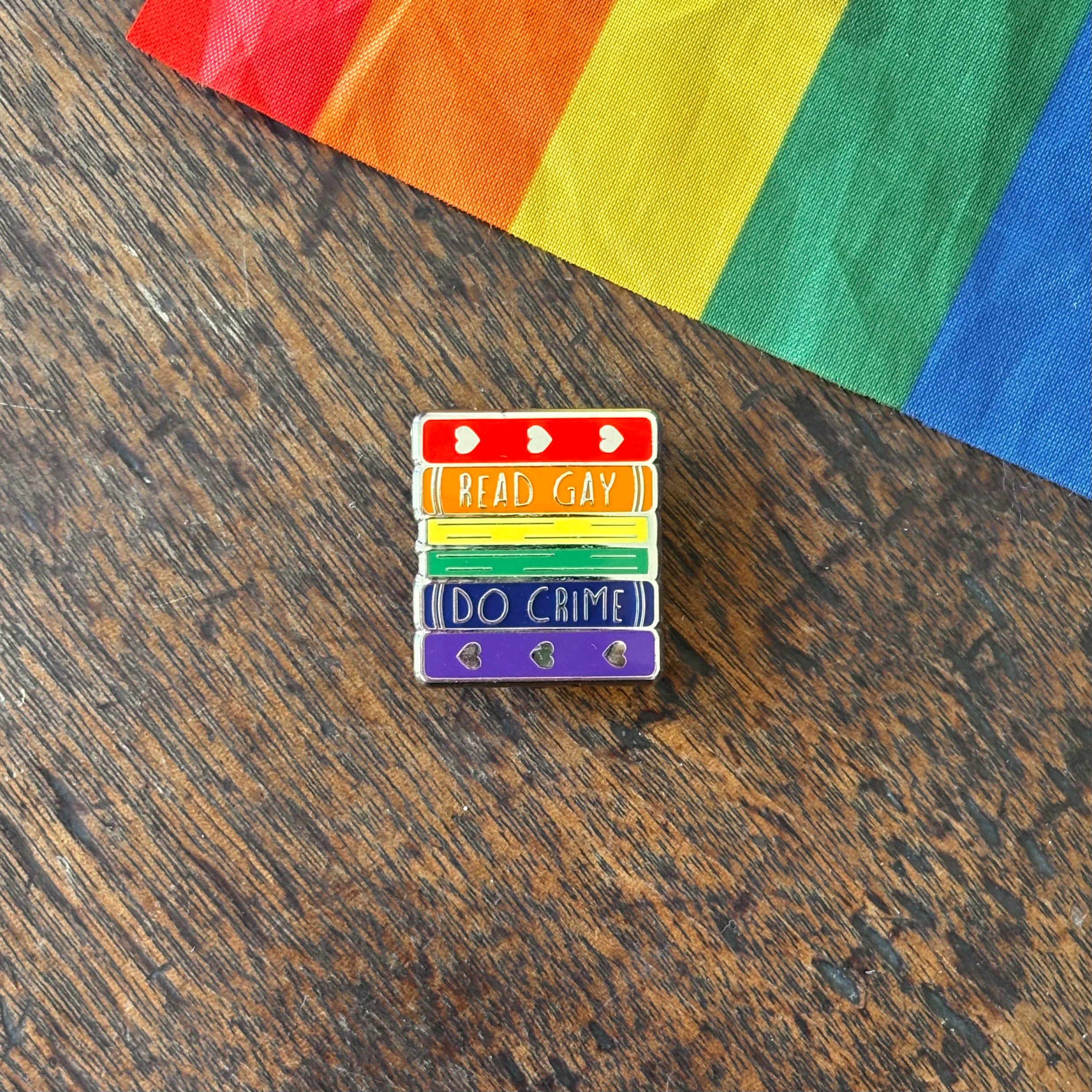Pin with 'Read Gay, Do Crime' text on a wooden surface with a rainbow fabric background