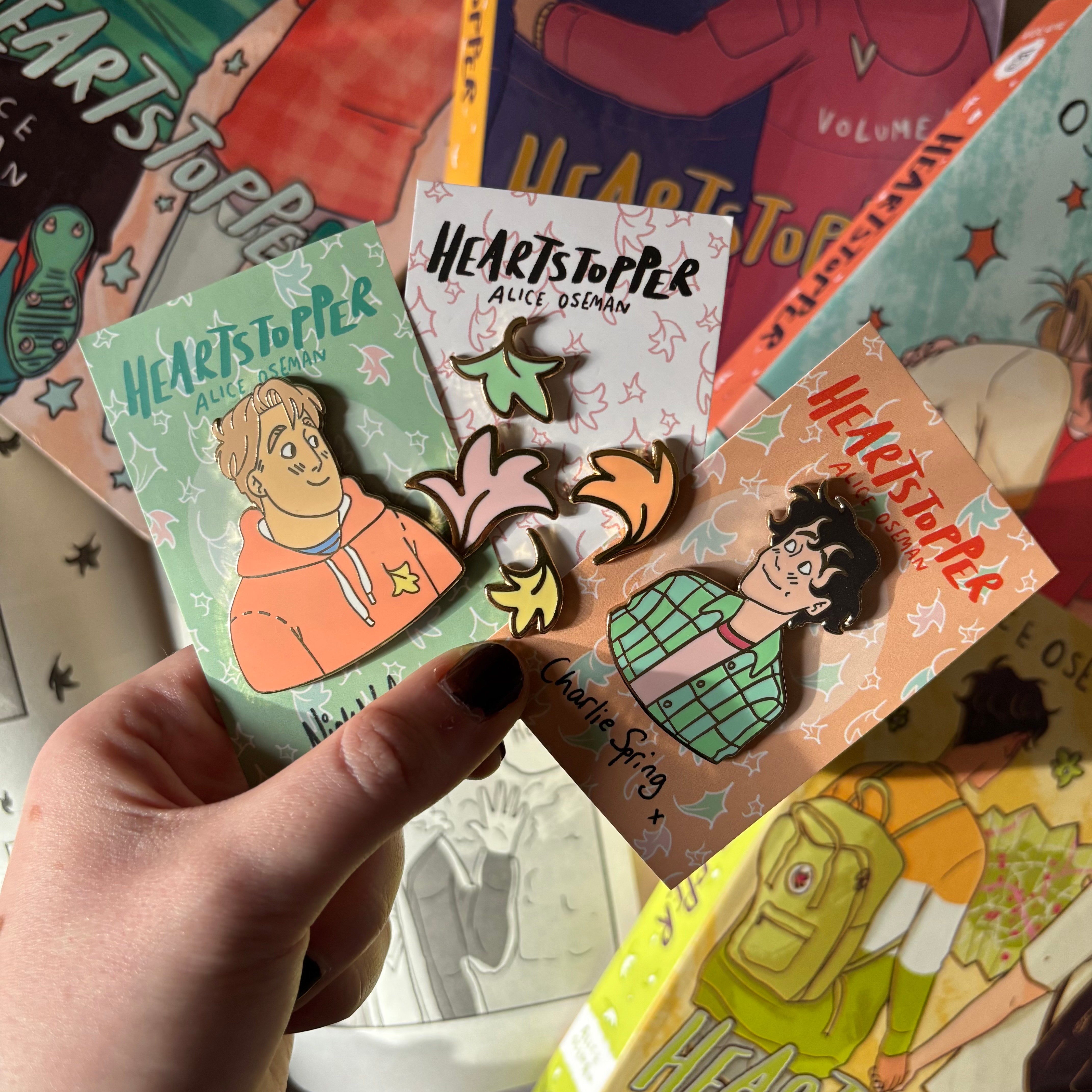 A bundle of our officially licensed Heartstopper pins. Features Nick Nelson, Charlie Sprint, and mini leaf pins, all designed by Alice Oseman. Held against a background of Heartstopper graphic novels.