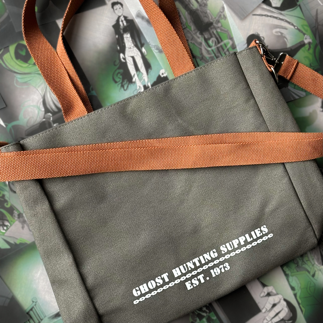 Grey canvas tote bag with white text reading "Ghost Hunting Supplies est. 1973" printed on the front. The bag has brown straps and is pictured against a background of page overlays.