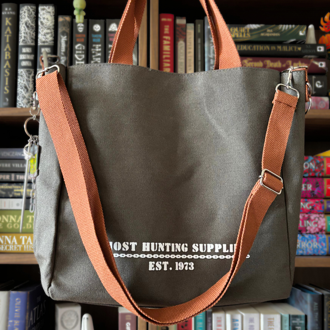 Grey canvas tote bag with white text reading "Ghost Hunting Supplies est. 1973" printed on the front. The bag has brown straps and is pictured held against a bookshelf.