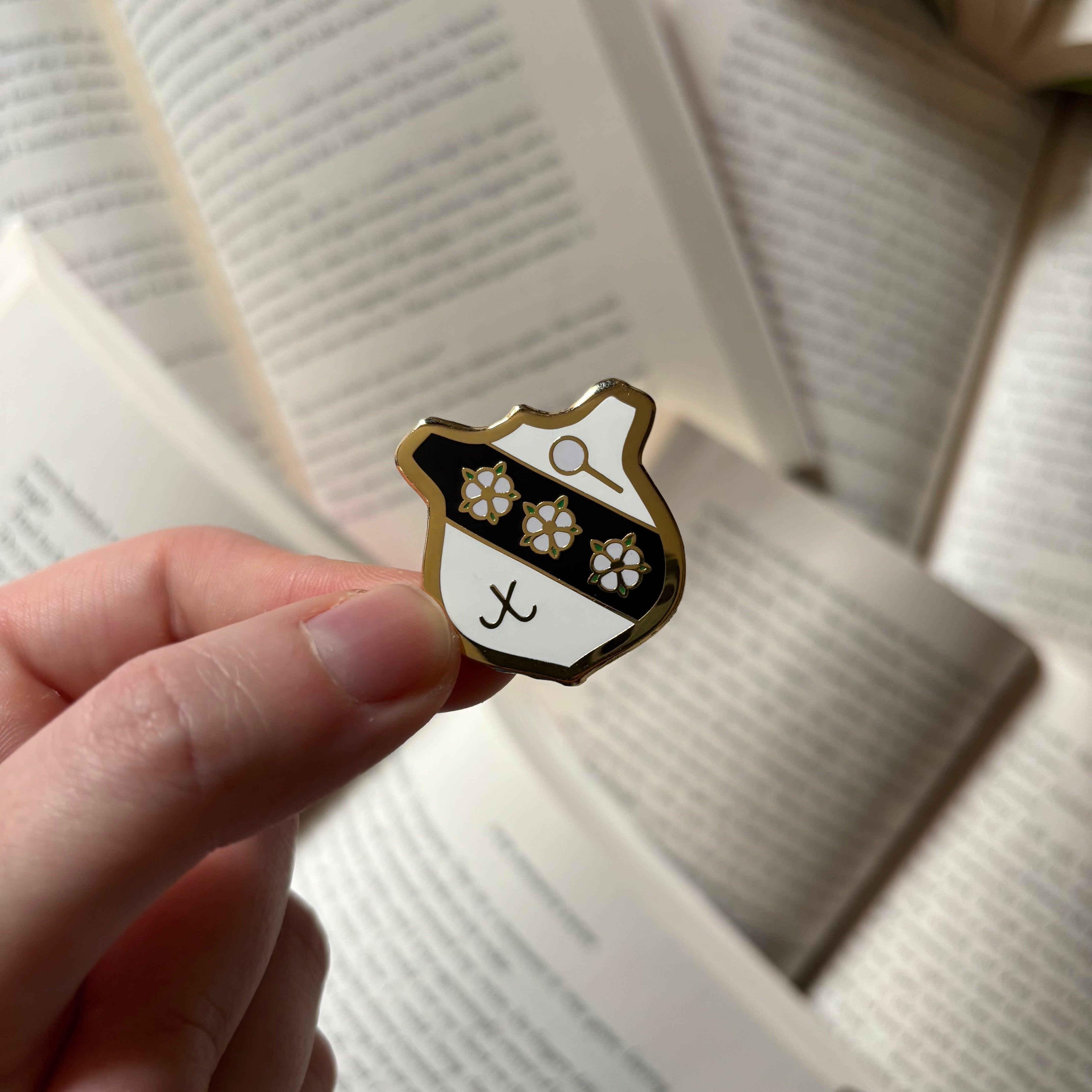 Official Murder Most Unladylike pin, featuring the crest of Deepdean school. Held above a background of blurred books