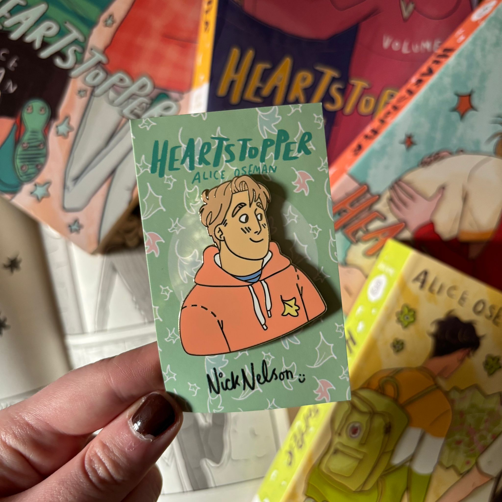 The Nick Nelson officially licensed enamel pin on a Heartstopper backing card, being held above a blurred background of Heartstopper books by Alice Oseman.
