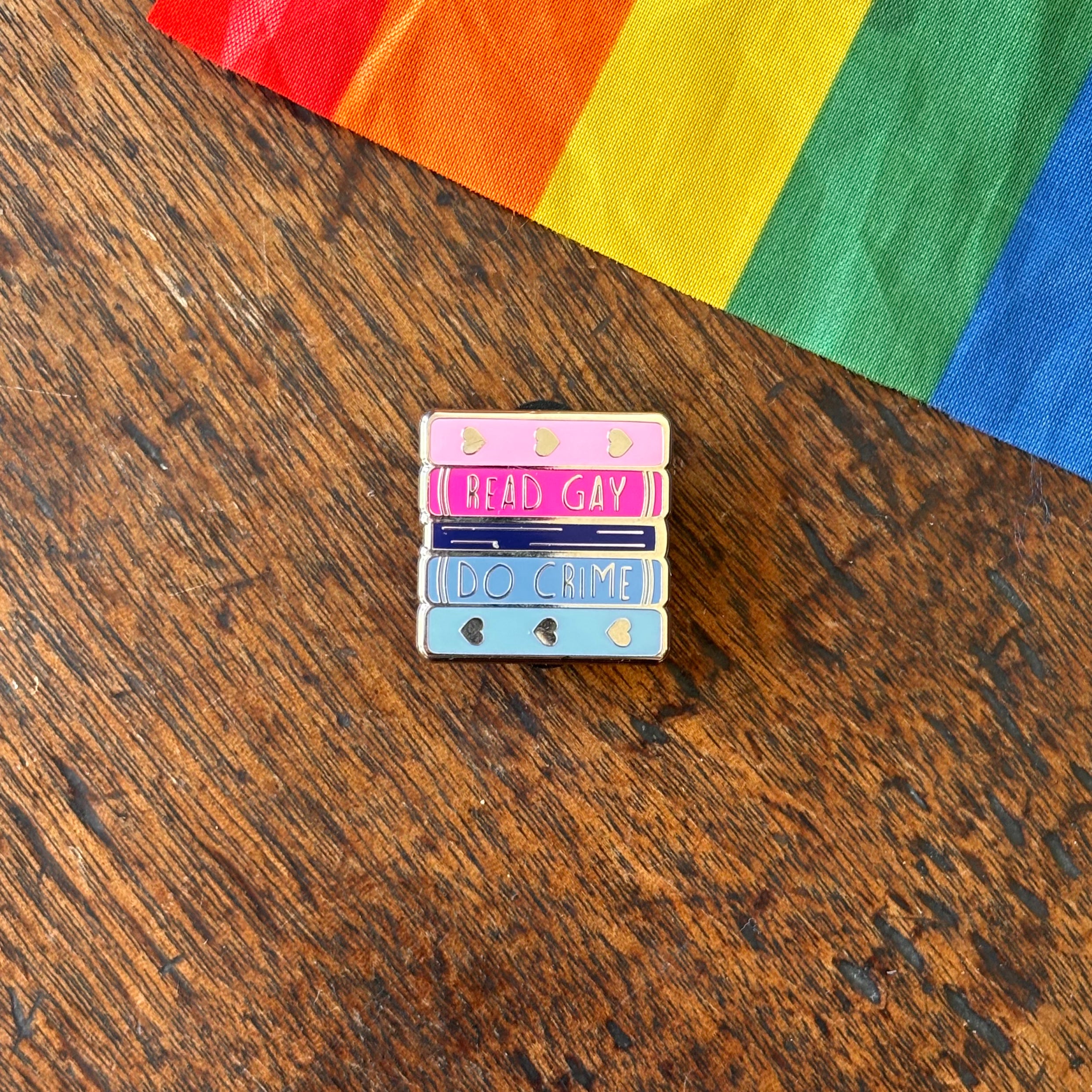 Pin with 'Read Gay, Do Crime' text on a wooden surface with a rainbow fabric background