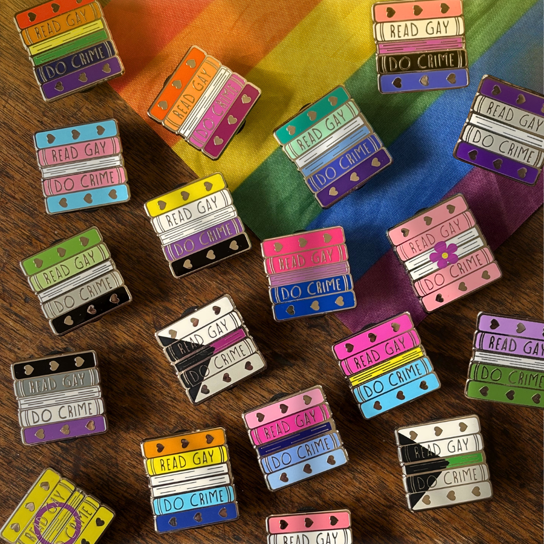 Enamel pins shaped like stacks of books, coloured in the colours of various pride flags. The pins feature the text "Read Gay Do Crime" and are pictured against a pride flag and wooden background.