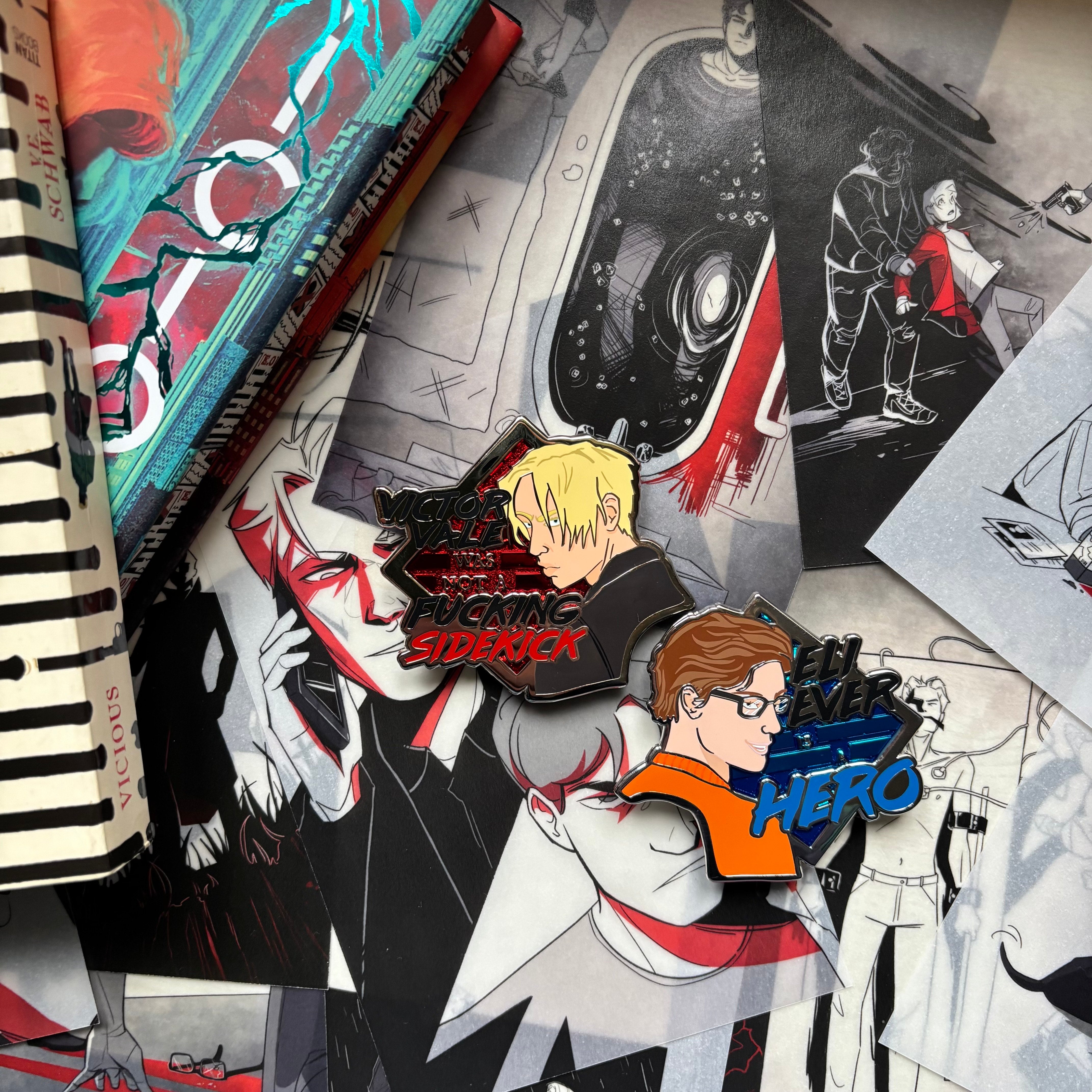 LitPins&Co Vicious Deluxe Enamel Pins resting against the page overlays. The pins feature Victor Vale and Eli Ever along with their iconic quotes: "Victor Vale was not a f**king sidekick" and "Eli Ever is a Hero"