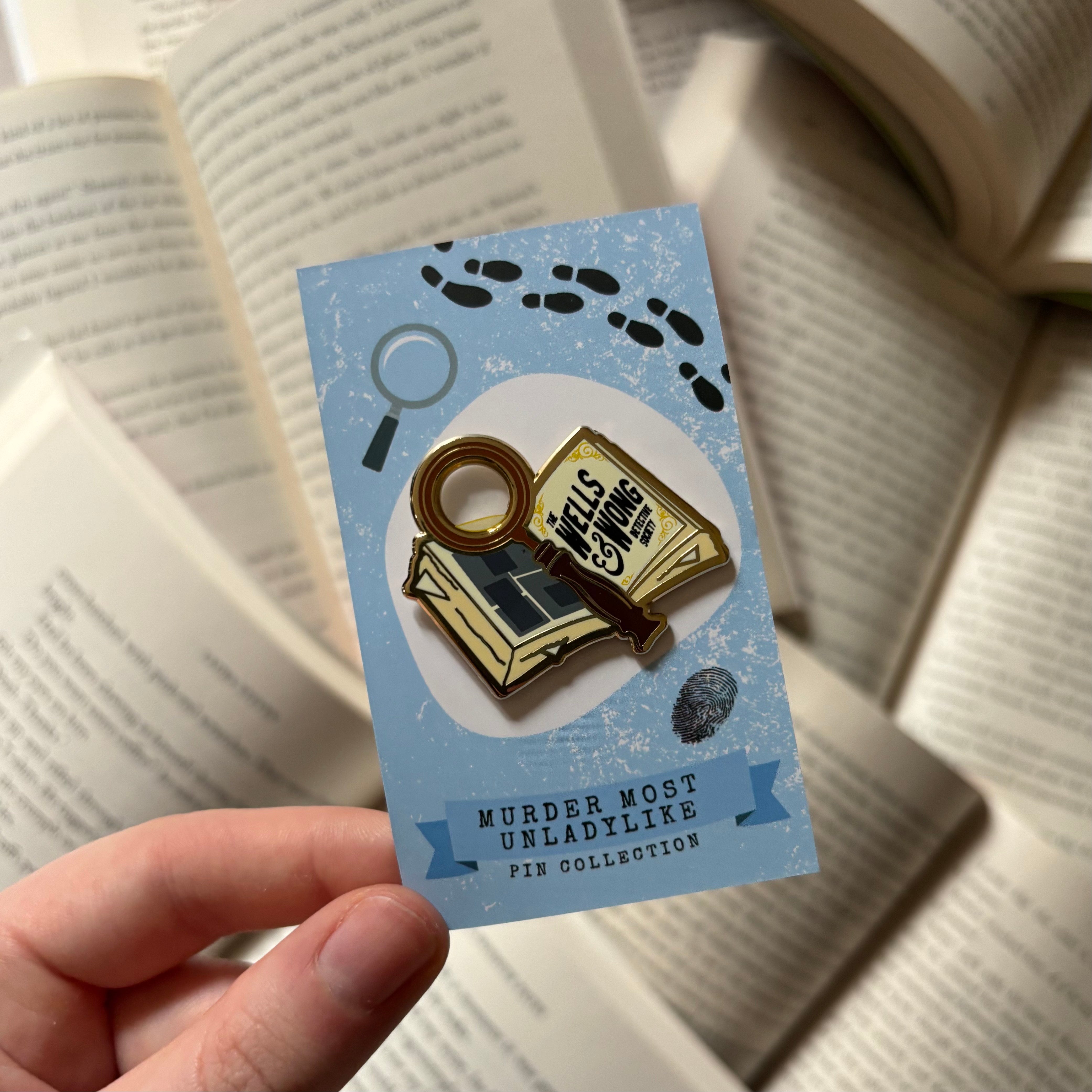 Officially licensed Murder Most Unladylike pin, featuring a book and a magnifying glass with the text "The Wells & Wong Detective Society," held against a background of blurred books.