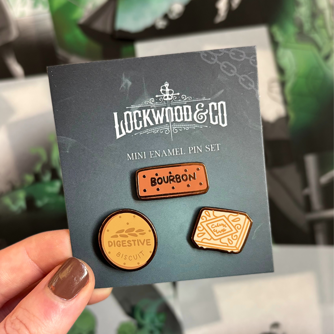 Set of mini biscuit enamel pins displayed on a Lockwood&Co backing card, held against a blurred background of page overlays.