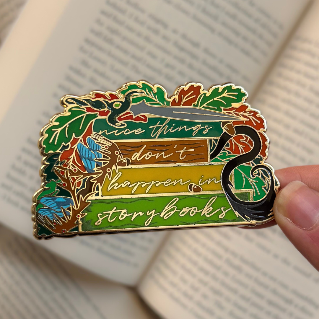 Enamel pin inspired by The Cruel Prince by Holly Black, reading "Nice things don't happen in storybooks." Held against a background of open books.