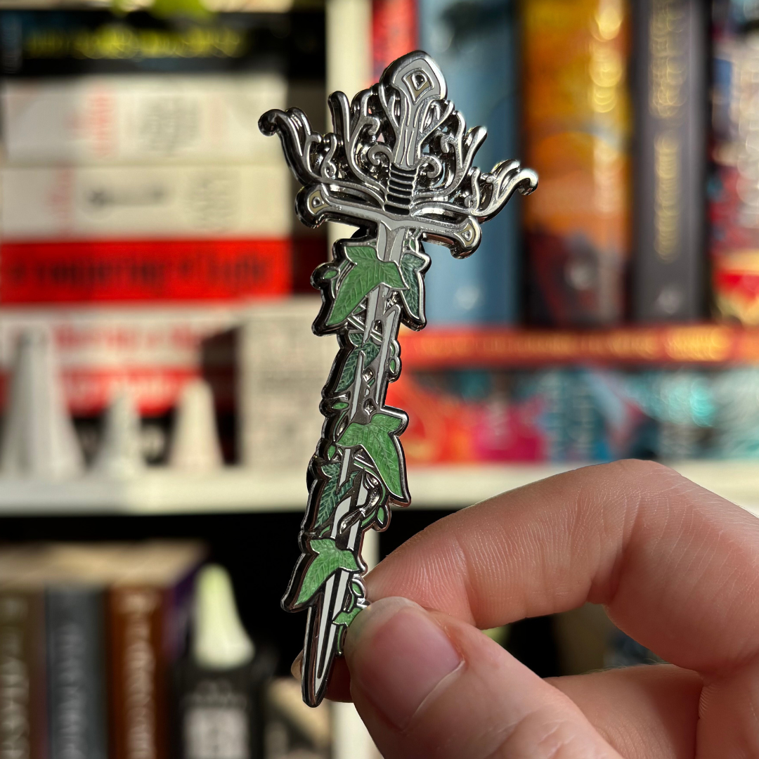 Sword pin inspired by the fantasy world of Middle Earth, held against a blurred background of bookshelves.