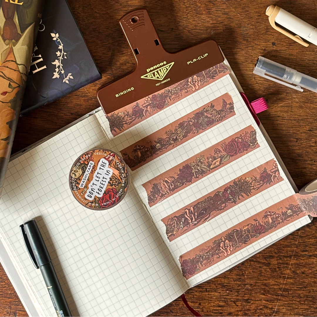 An open journal page decorated with brown washi tape. The washi tape features a dark botanical design of flowers, bones, mushrooms, leaves, and more.