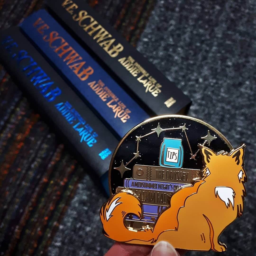 Books Feed The Mind, Tips Feed The Cat enamel pin above three different hardback copies of The Invisible Life of Addie LaRue by V.E Schwab