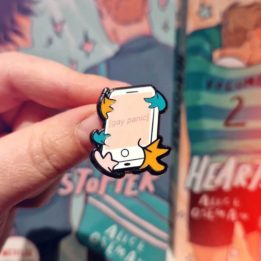 Heartstopper inspired enamel pin with 'Gay Panic' design, held in front of the Heartstopper graphic novels by Alice Oseman.