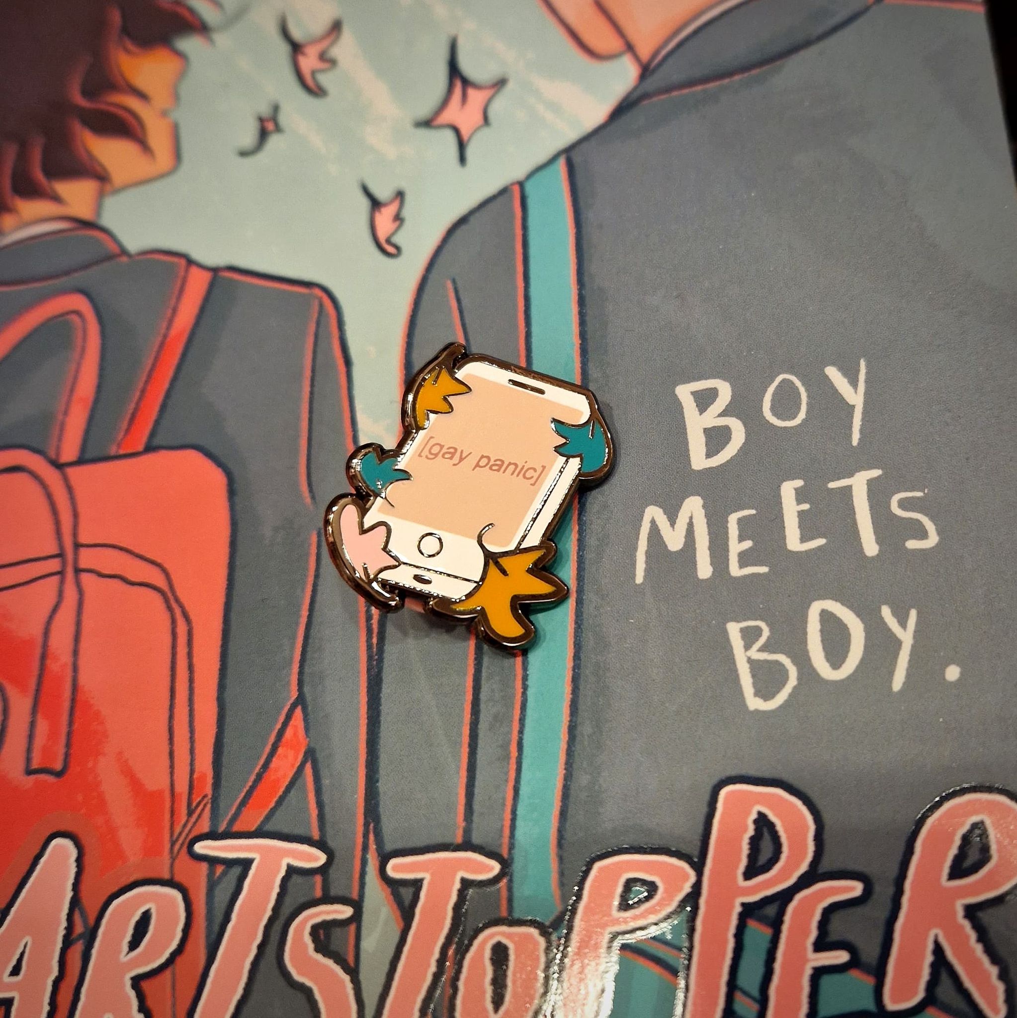 Heartstopper inspired enamel pin with 'Gay Panic' design, held in front of the Heartstopper graphic novels by Alice Oseman.