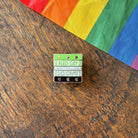 Pin with 'Read Gay, Do Crime' text on a wooden surface with a rainbow fabric background