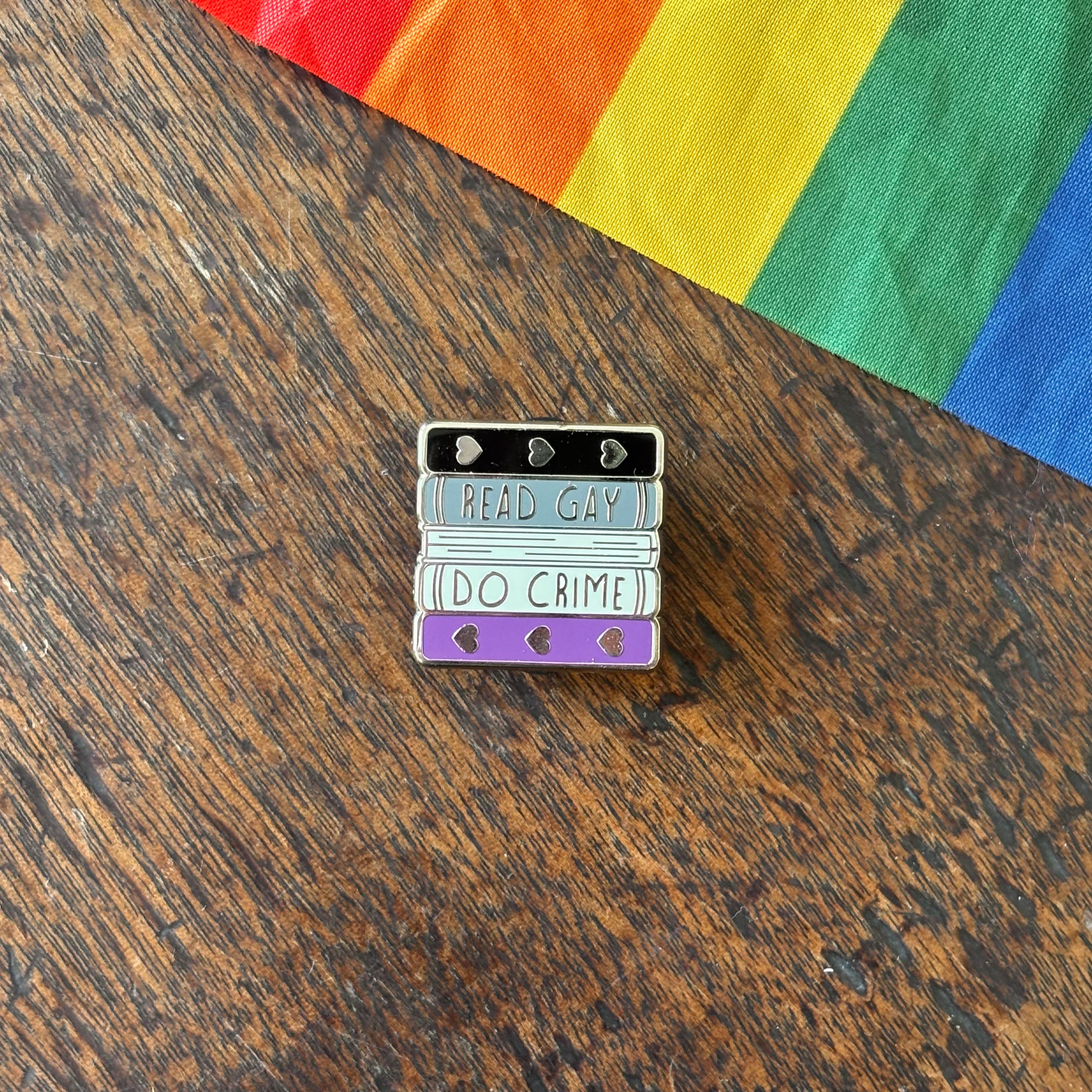 Pin with 'Read Gay, Do Crime' text on a wooden surface with a rainbow fabric background