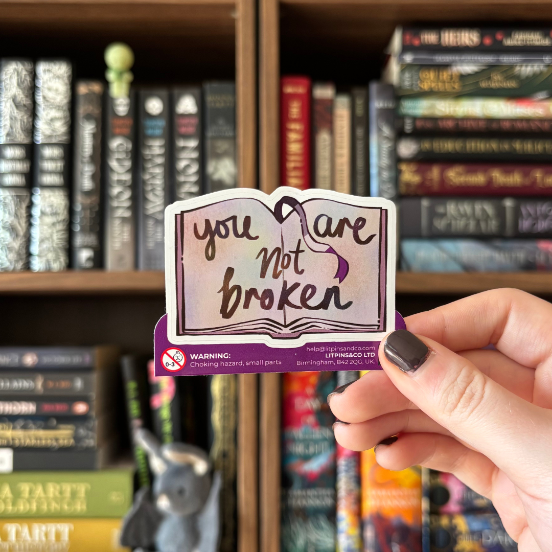 Hand holding a 'you are not broken' asexual themed book sticker in front of a bookshelf