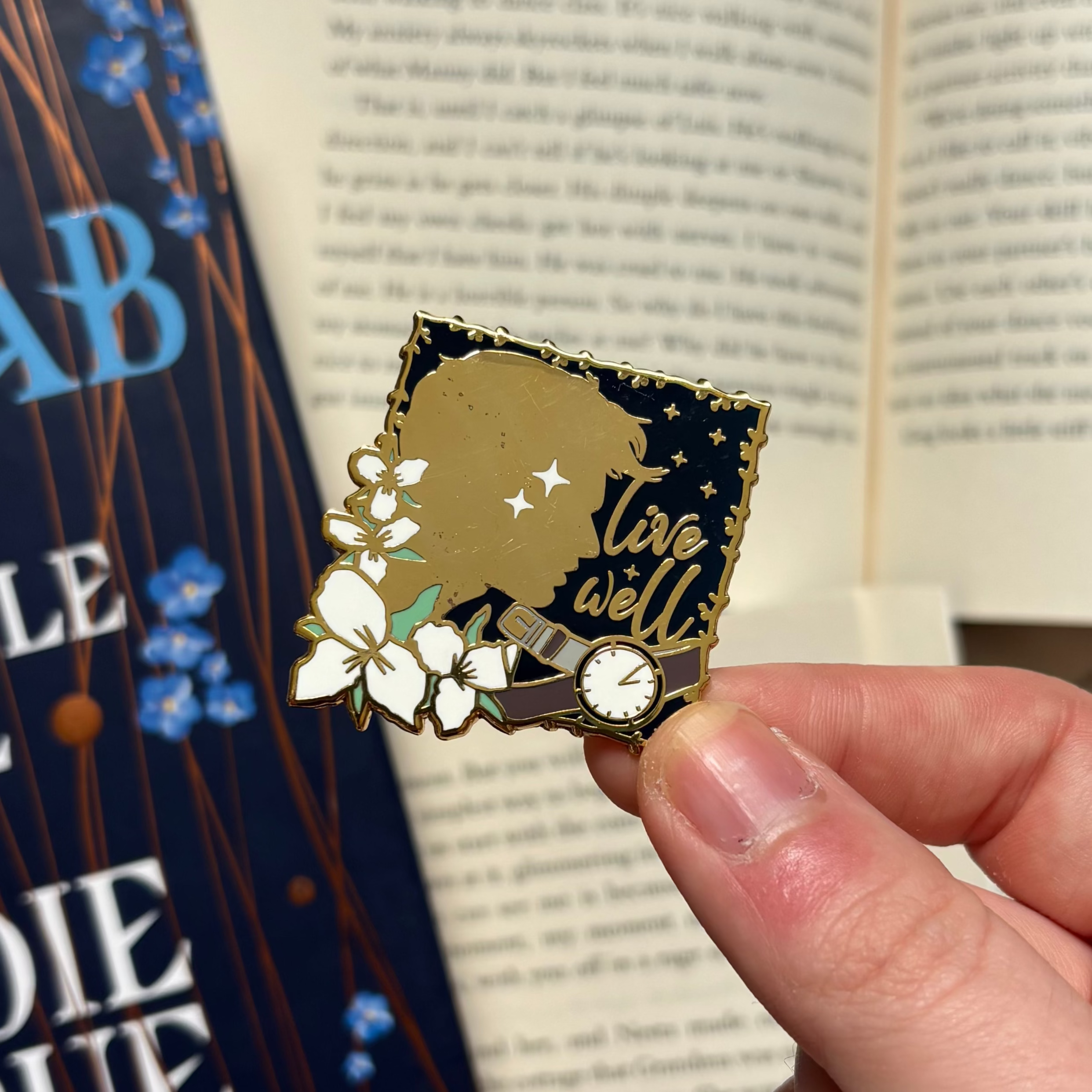 Pin with silhouette, flowers, and 'live well' text inspired by The Invisible Life of Addie LaRue, held in front of a background of books