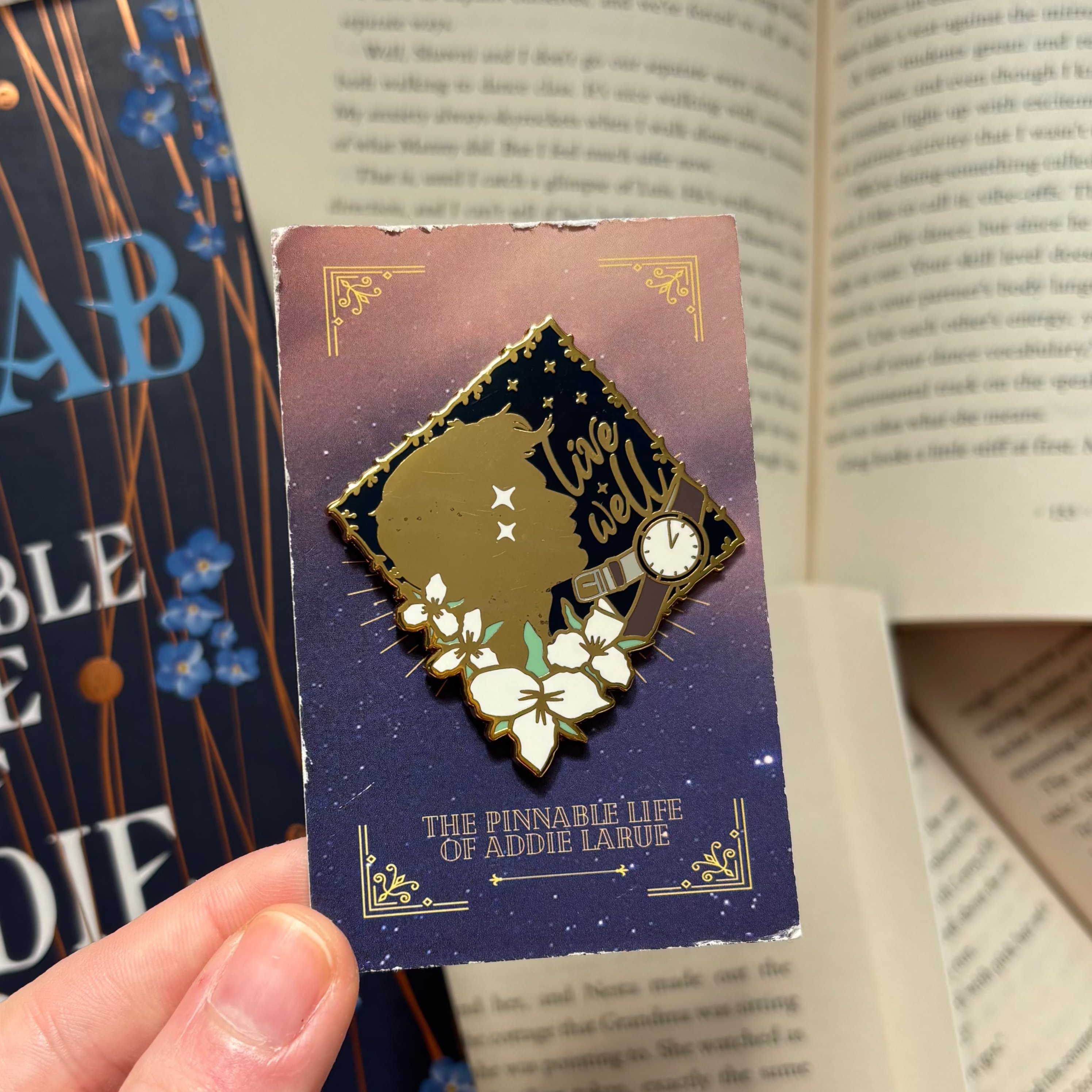 Pin with silhouette, flowers, and 'live well' text inspired by The Invisible Life of Addie LaRue, held in front of a background of books