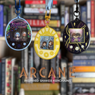 Three Tamagotchi style shaker keychains inspired by TV show Arcane, featuring characters Jayce, Viktor, Jinx, Ekko, and Powder. Held against a bookshelf.