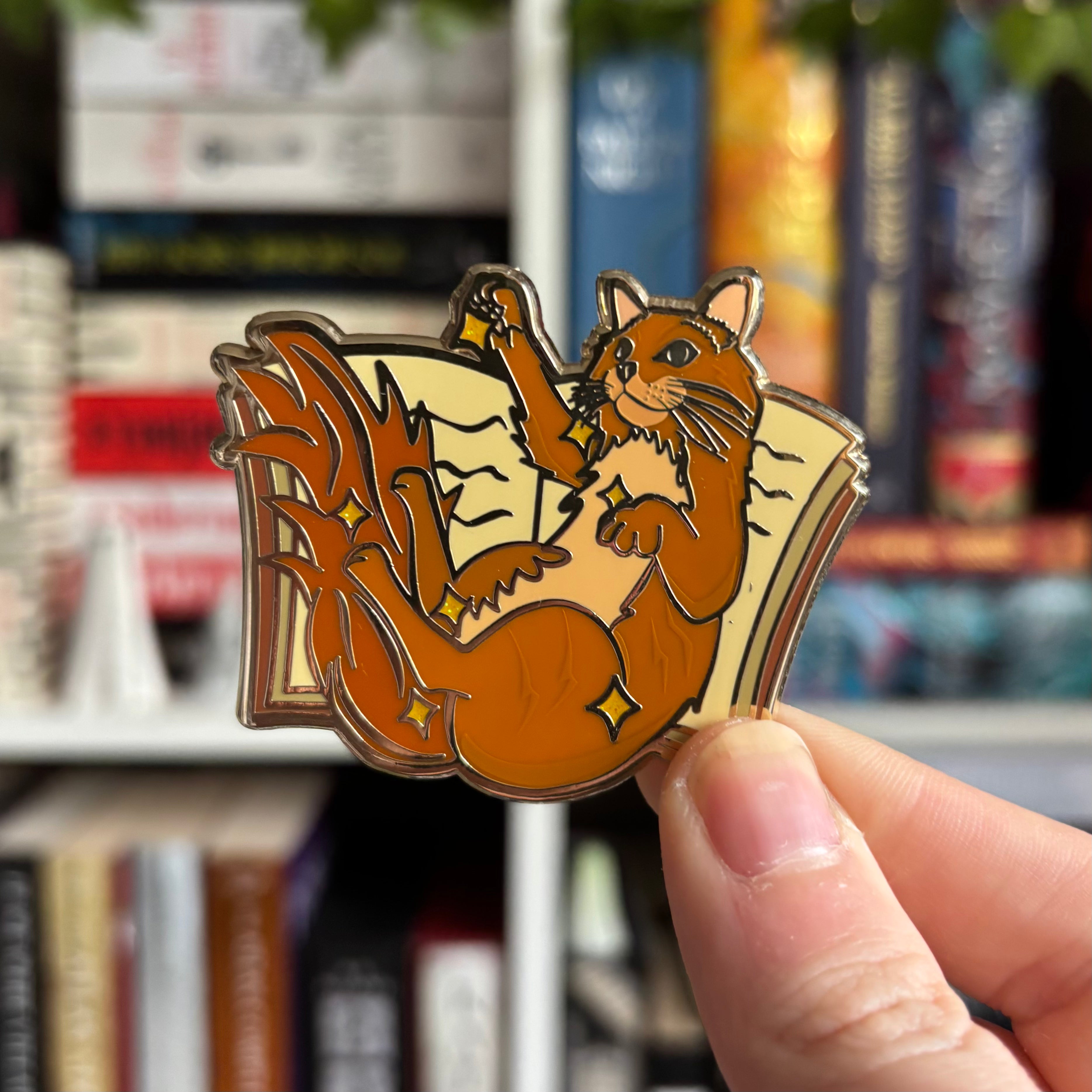 Enamel pin inspired by Book the cat from V. E. Schwab's Addie LaRue, featuring a cat pictured against an open book. Pin is held against a blurred background of books.