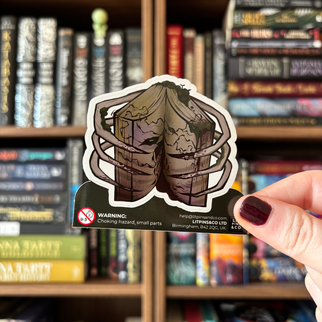 A die-cut sticker featuring a cracked, bloody, and moss-ridden book held open by skeletal hands. The sticker is held against a blurred bookshelf.