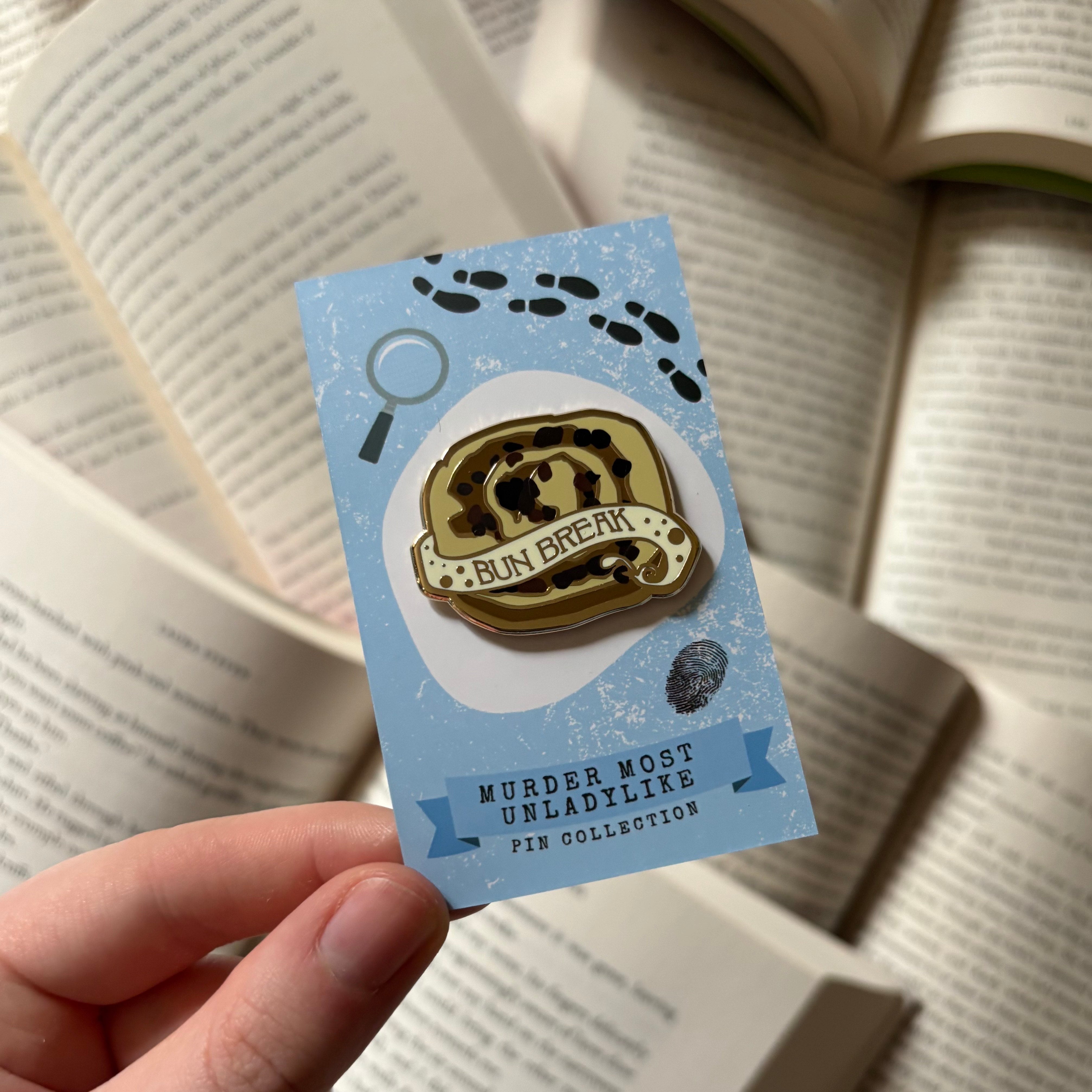 An enamel pin shaped like a bun, with the text "bun break," held above a background of open books