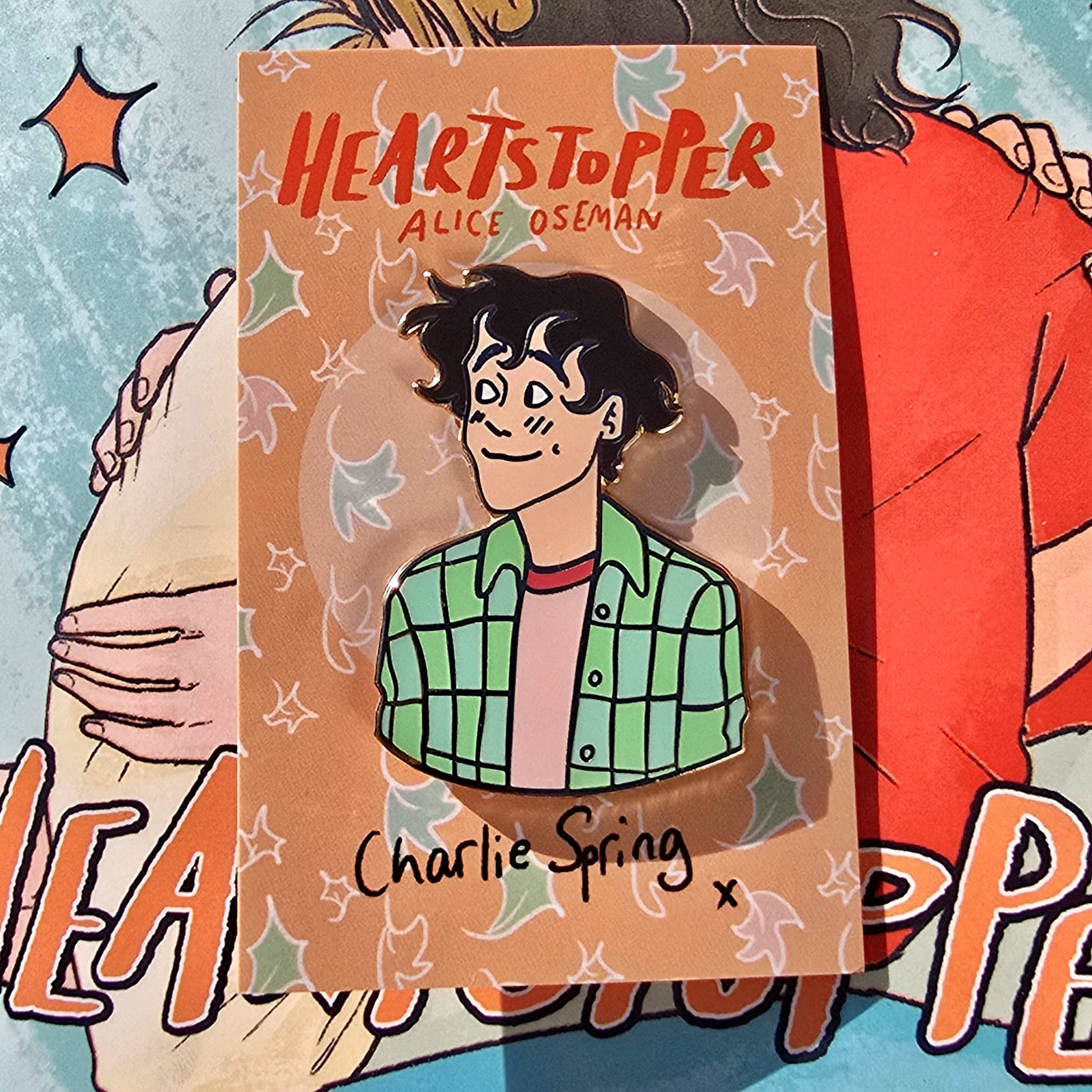 The Officially Licenced Heartstopper Charlie Sping Enamel Pin with backing card resting against a copy of Heartstopper by Alice Oseman.