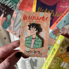 Alice Oseman's Heartstopper Charlie Spring enamel pin and backing card being held up against a background of blurred Heartstopper books.