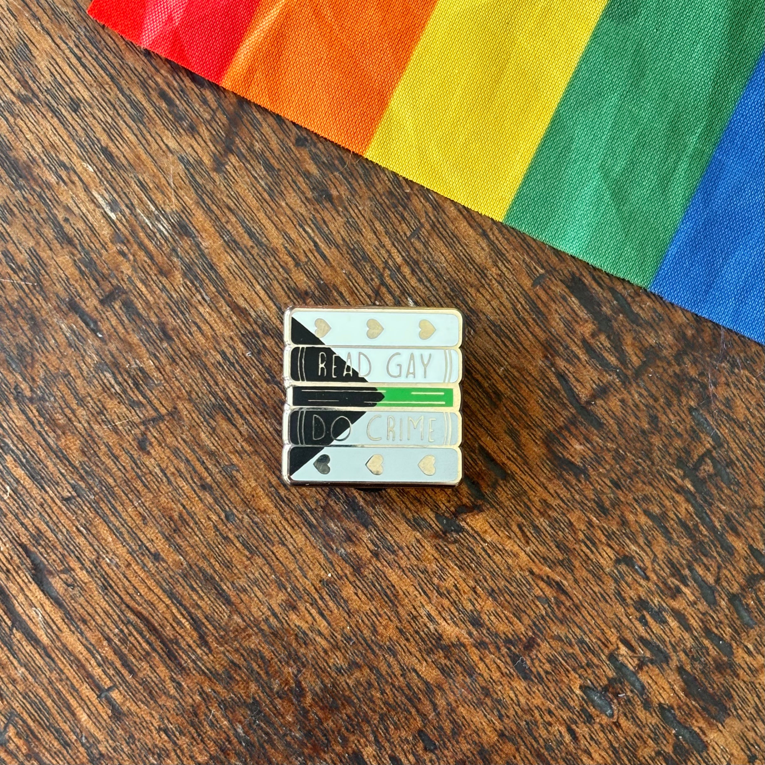 Pin with 'Read Gay, Do Crime' text on a wooden surface with a rainbow fabric background