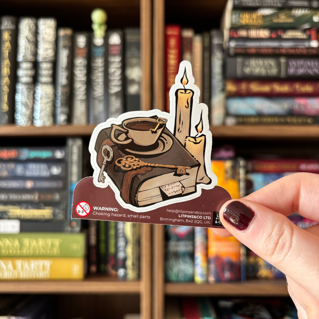 A hand with dark burgundy nail polish holds a die-cut sticker featuring an illustration of a brown leather-bound book, a cup of tea on a saucer, a key, and two lit candles. The sticker is positioned in front of a blurred bookshelf background.