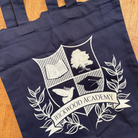A navy blue canvas tote bag with a white printed crest and the words 'WICKWOOD ACADEMY' laid flat on a wooden floor.