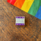 Pin with 'Read Gay, Do Crime' text on a wooden surface with a rainbow fabric background