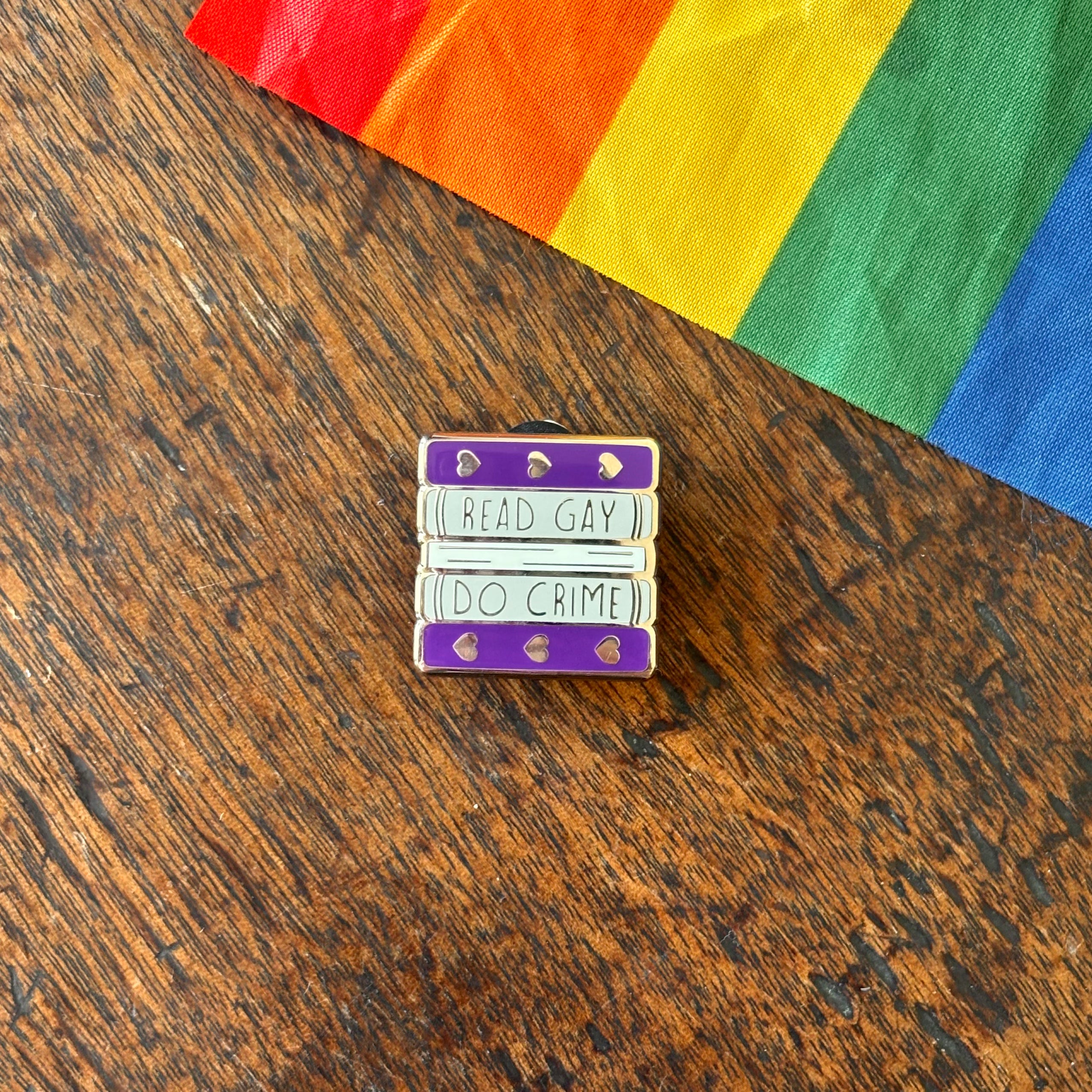 Pin with 'Read Gay, Do Crime' text on a wooden surface with a rainbow fabric background