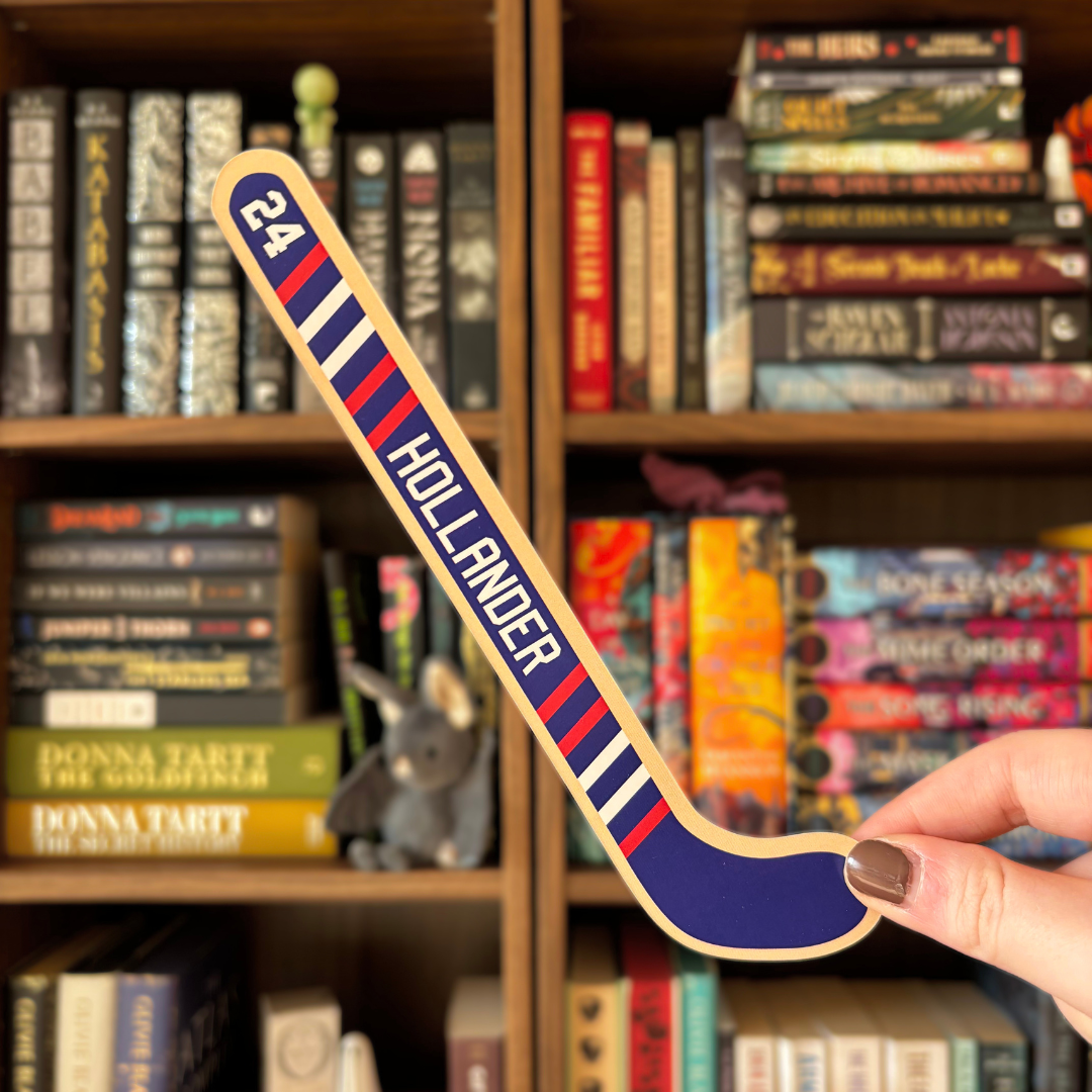 A blue bookmark shaped like a hockey stick with text reading "Hollander". The bookmark is held in front of a blurred bookshelf 